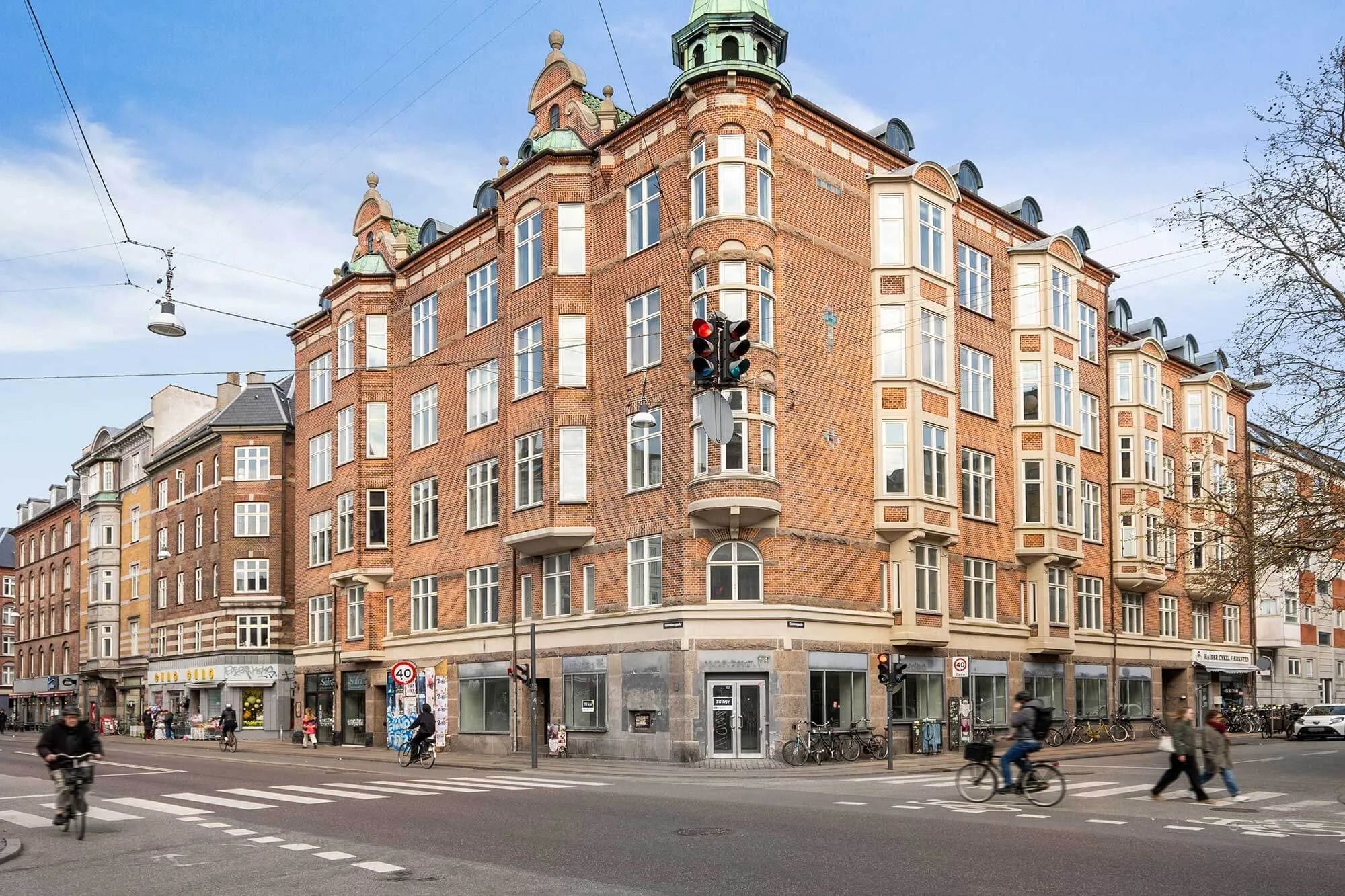 Mennesker cykler og går på en travl gade foran en stor, rødstenebygning. I baggrunden er der røde bygninger og trafiklys. På