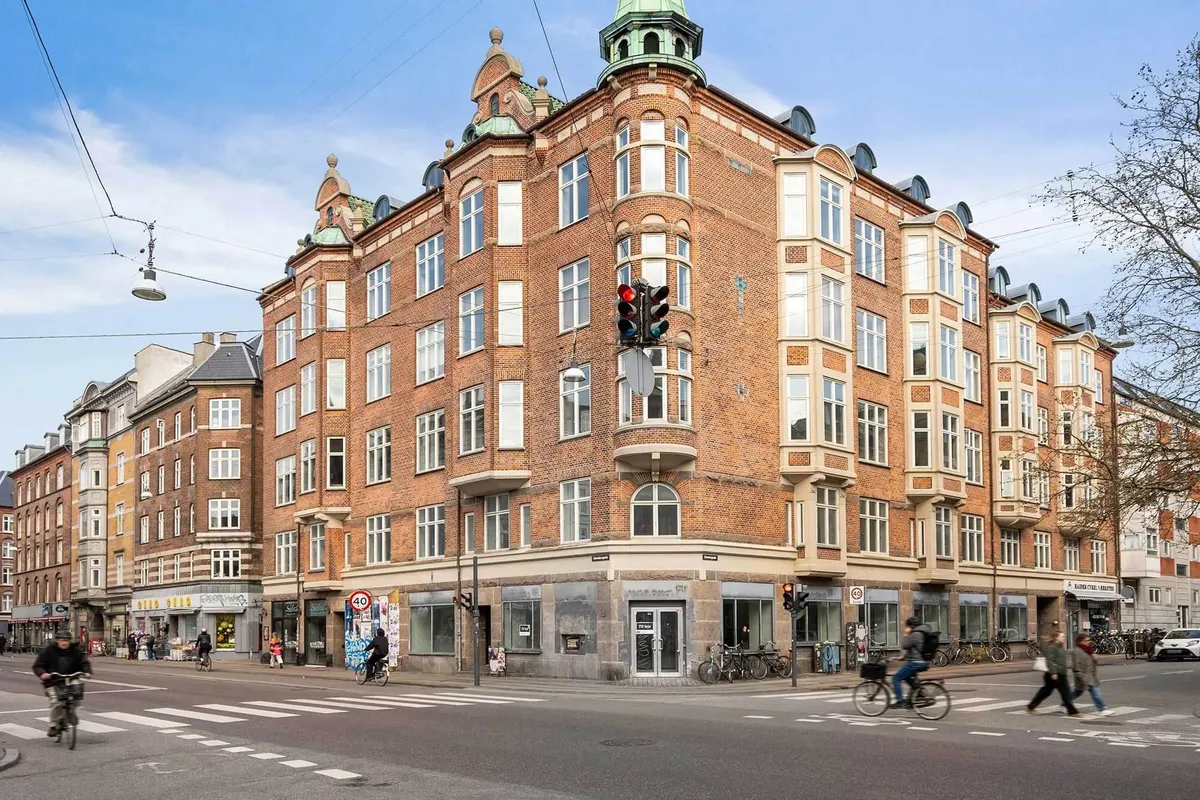 Mennesker cykler og går på en travl gade foran en stor, rødstenebygning. I baggrunden er der røde bygninger og trafiklys. På