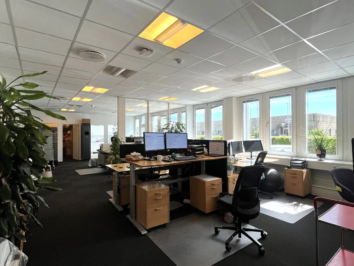 Kontor med mødelokale og lager, hvor flere skriveborde og computere står i tæt opstilling. Vægge er hvide, og der er lyse