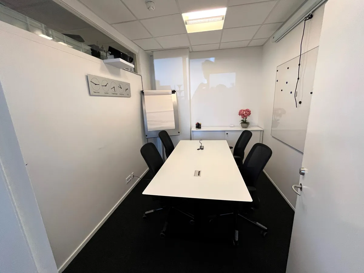 Mødelokale med 6 stole, hvide vægge og gulv og et bord med en vase med røde blomster.