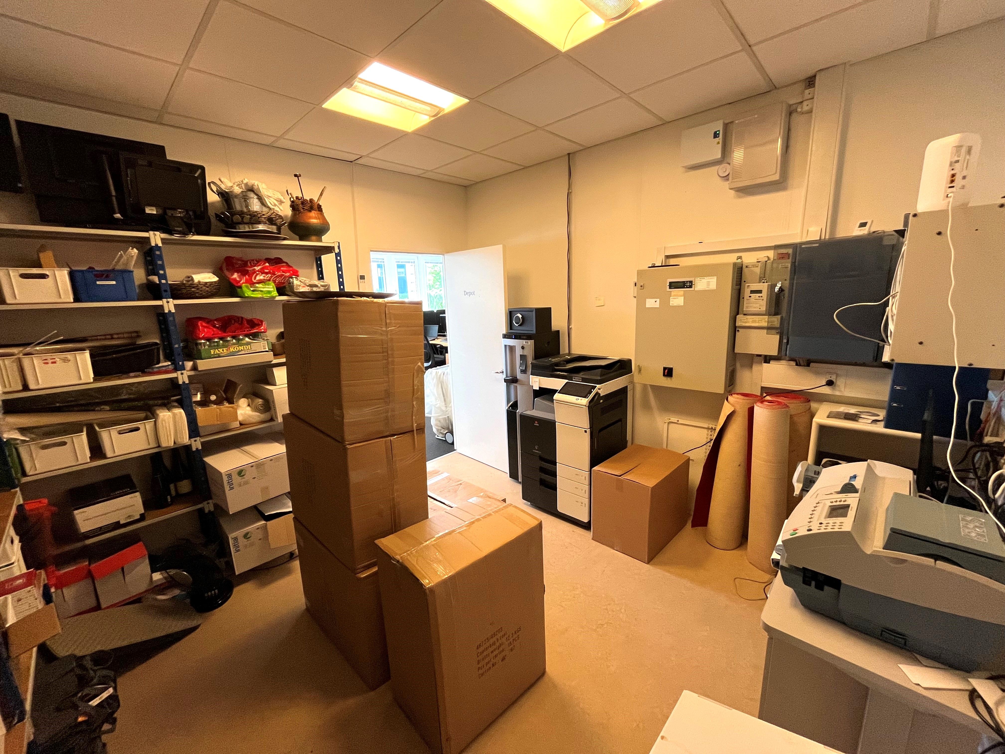 En lager- og mødelokale med flere kartoner, printere og skraldespande samt et skrivebord med en fax.