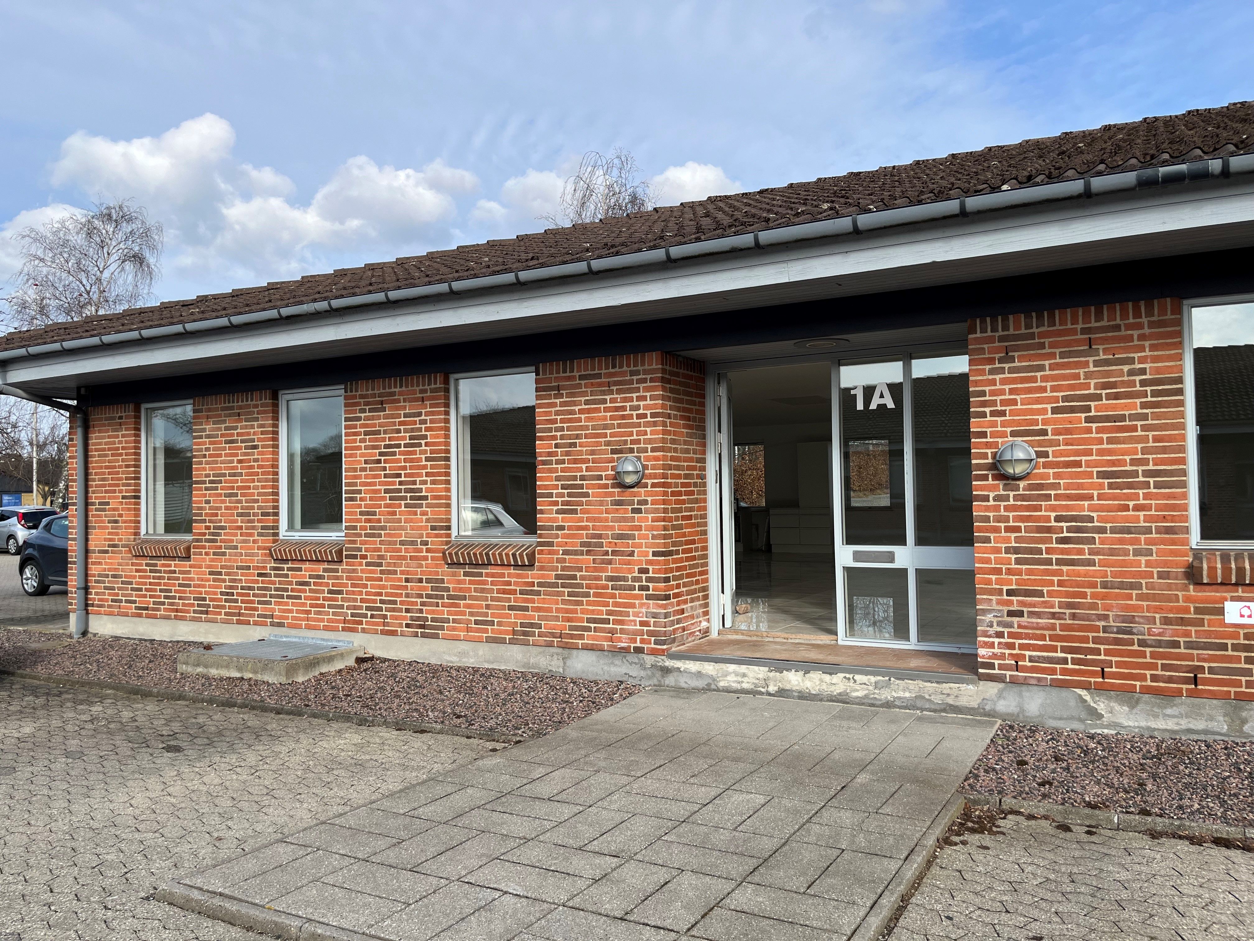 En lejlighed med glasvinduer og en indgang med nummer 1A, parkeringsplads med biler og et tage med mos og tagrør