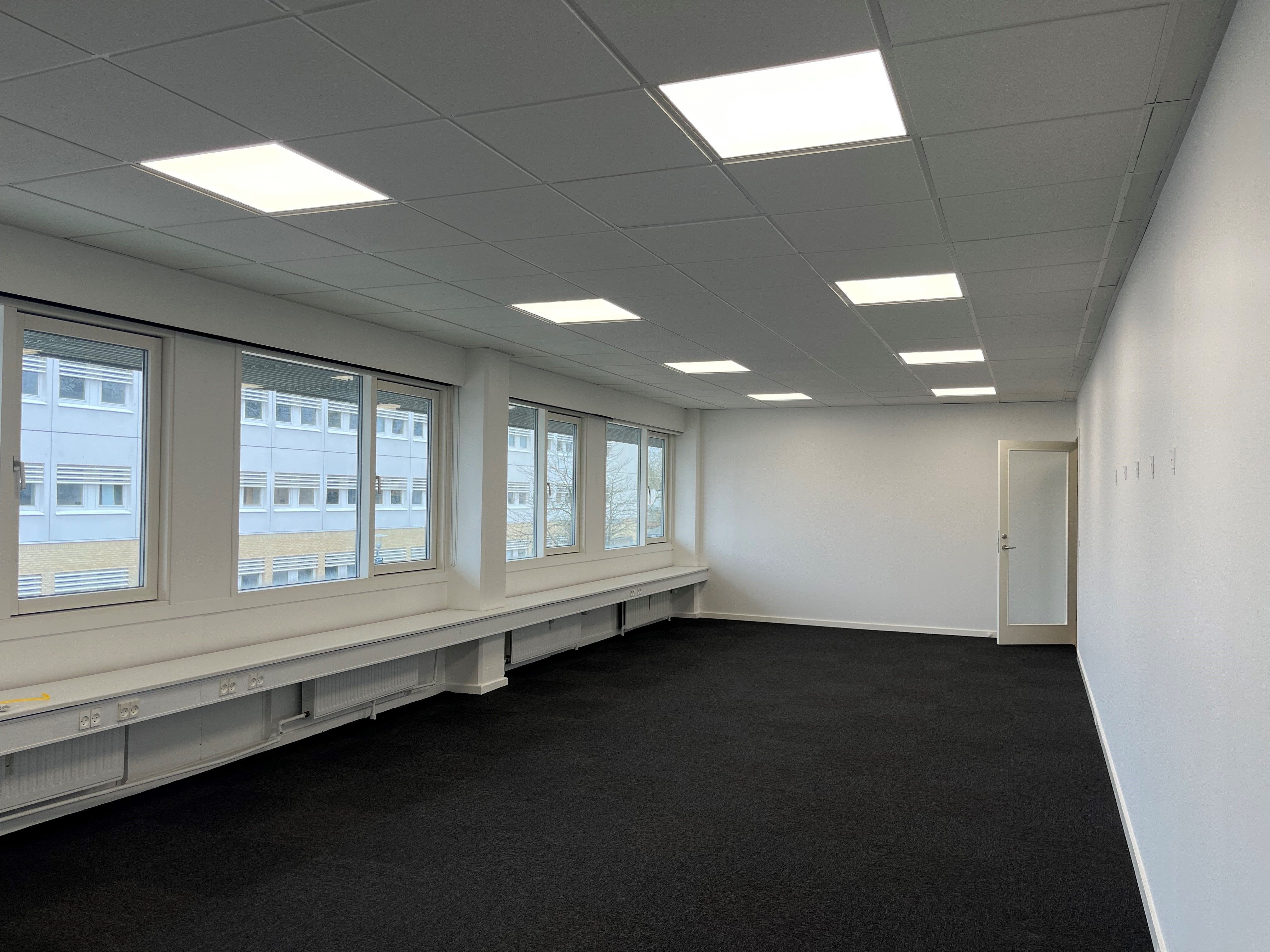 Et tomt kontor med mørke gulvbelægning, hvide vægge og vinduer med glas og hvide rammer, der vender ud mod en bygning med