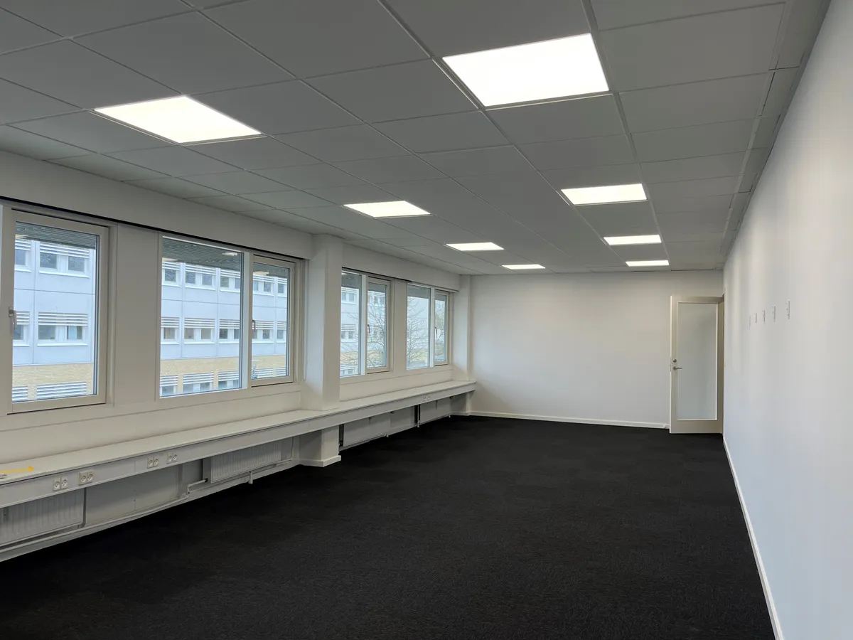 Et tomt kontor med mørke gulvbelægning, hvide vægge og vinduer med glas og hvide rammer, der vender ud mod en bygning med