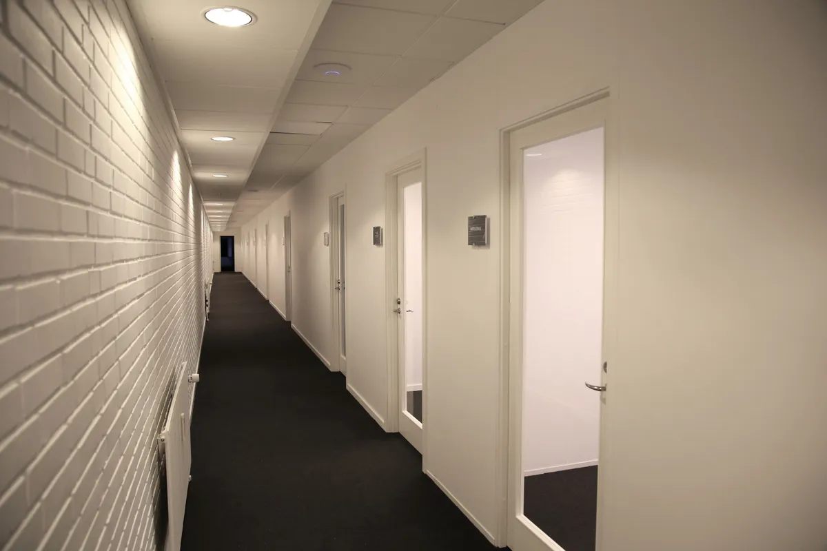 En lang hal med hvide vægge og hvide døre samt en sort gulvbelægning. Der er et lys i loftet, og der er flere døre til