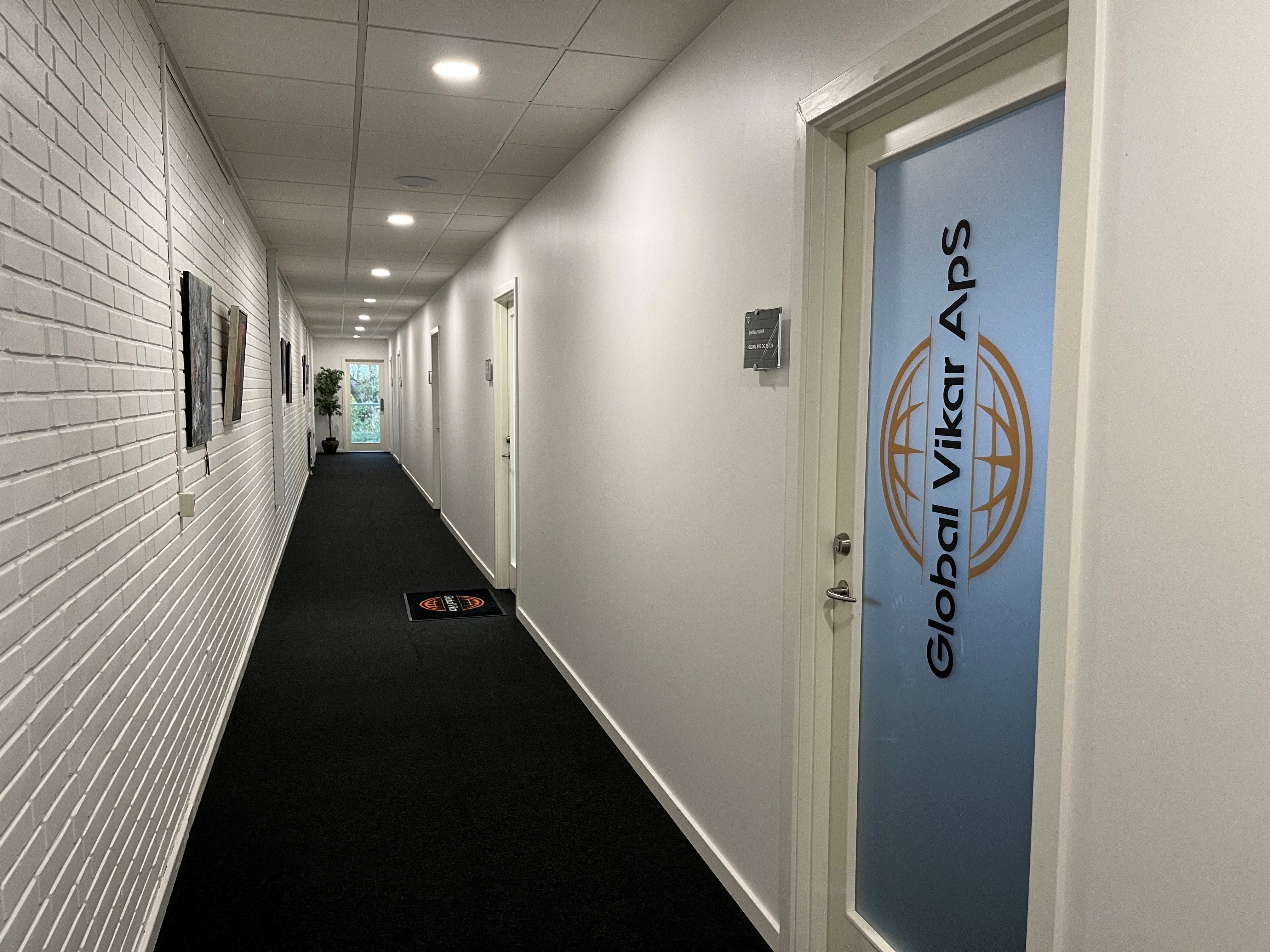 Kort korridor med sort gulvtæppe og hvide vægge, to døre og en plante i det fjerne