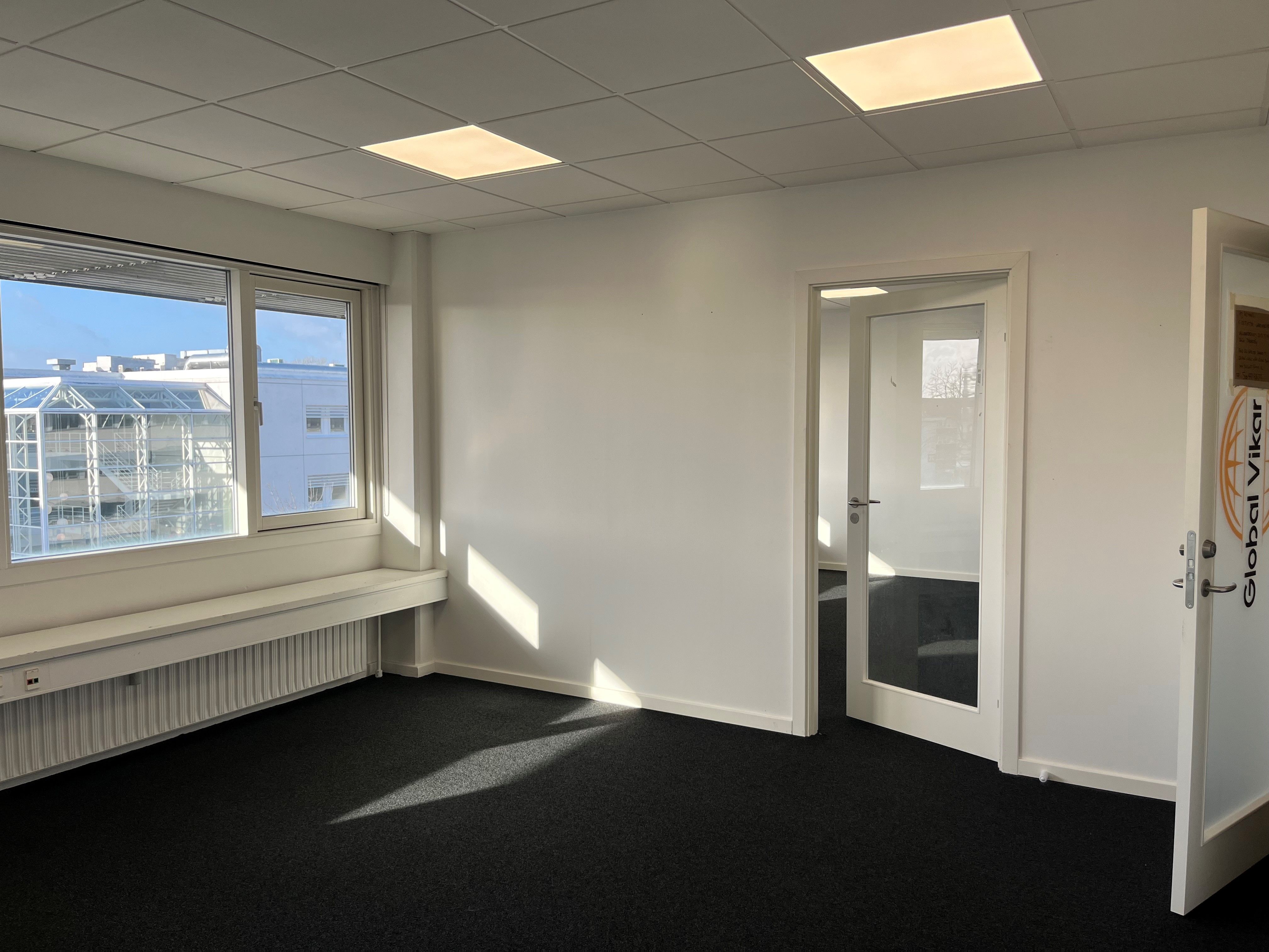 Et tomt kontor med to døre og vinduer med udsigt til et glashus. Et radiator er installeret under vinduet. Der er et