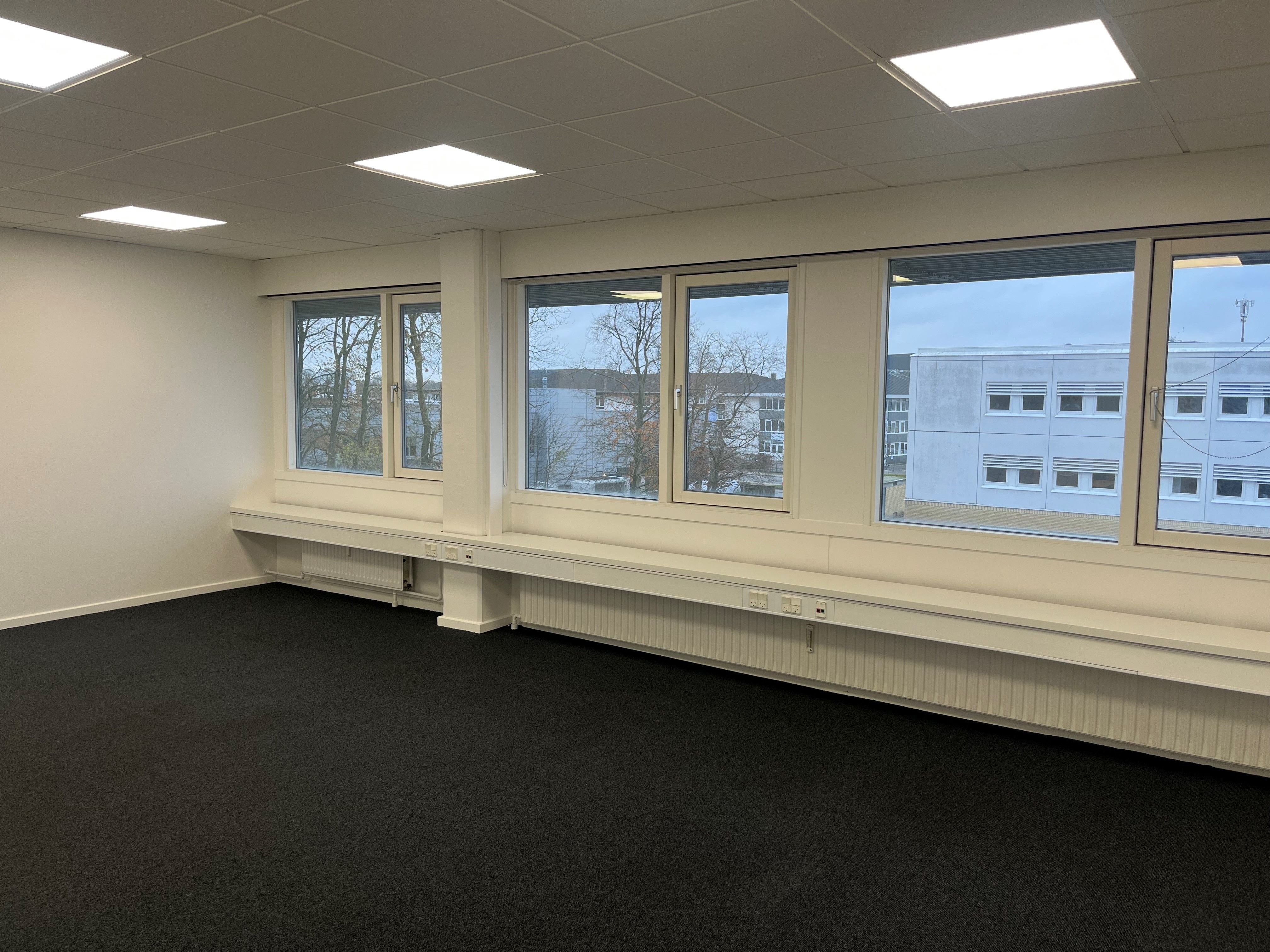 Store lyse vinduerne med udsigt til bygninger og træer. Hvide vægge og loft med indlejrede lys. Sorte gulvbelægning og hvide