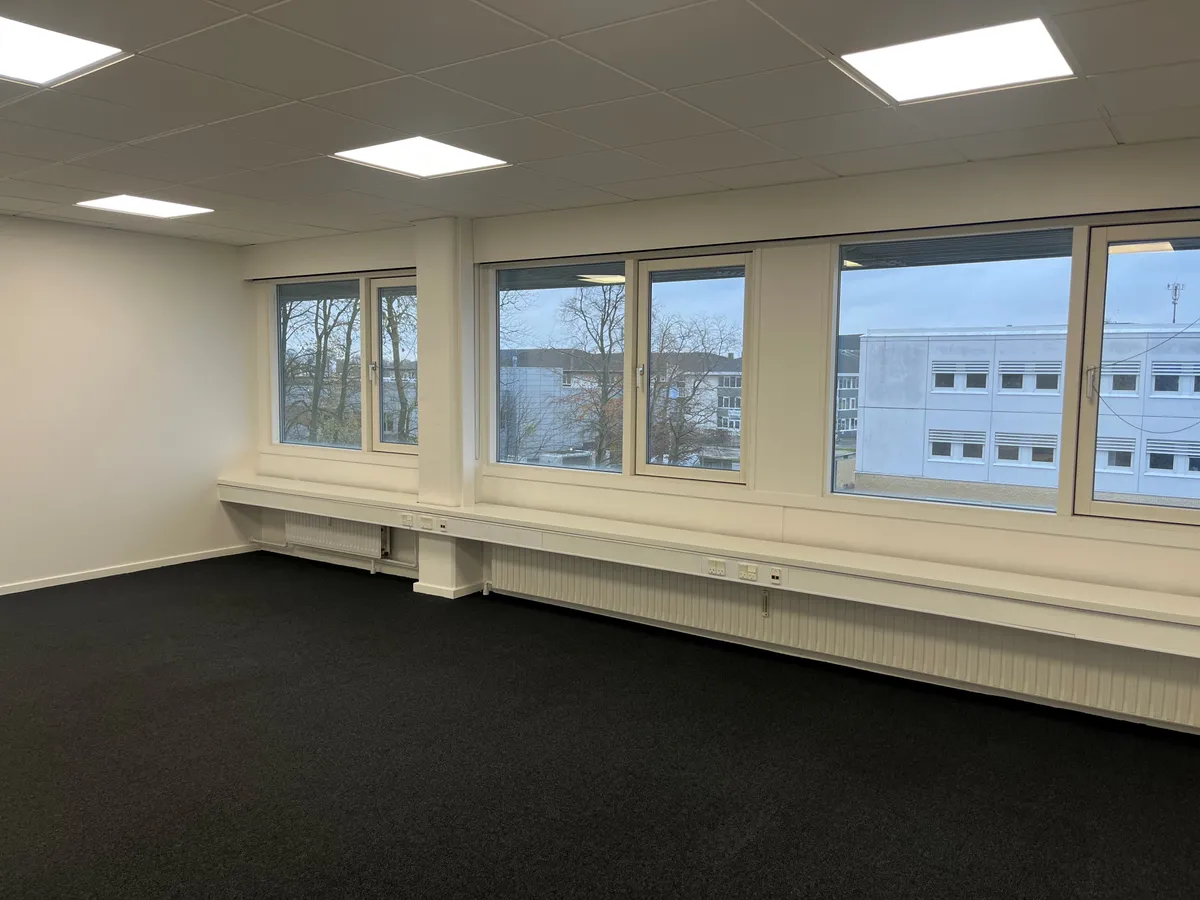 Store lyse vinduerne med udsigt til bygninger og træer. Hvide vægge og loft med indlejrede lys. Sorte gulvbelægning og hvide