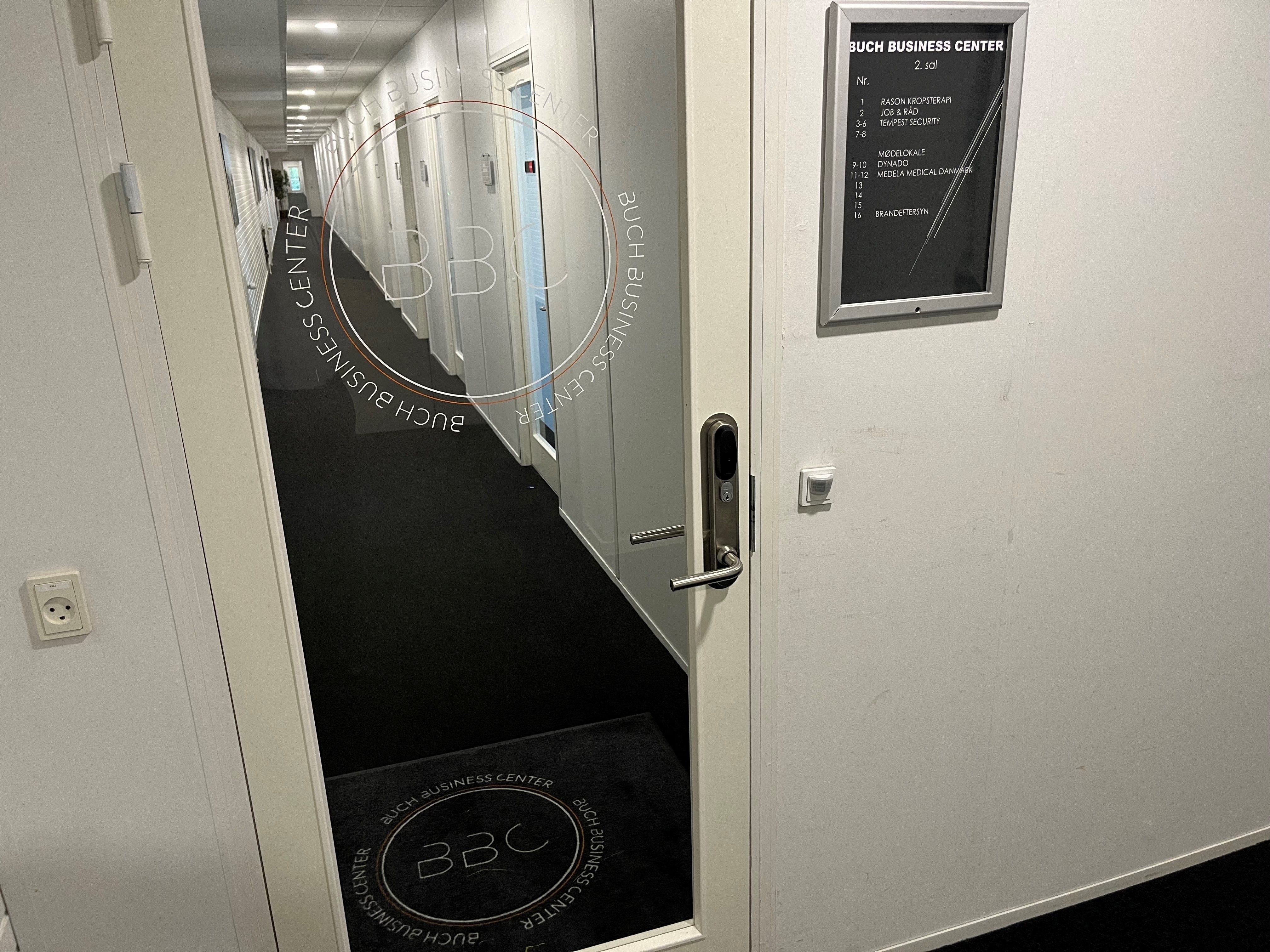 Gangen i et erhvervscenter med teksten "BUCH BUSINESS CENTER" og en dør med logoet "BBC" og elektriske stikkontakter på