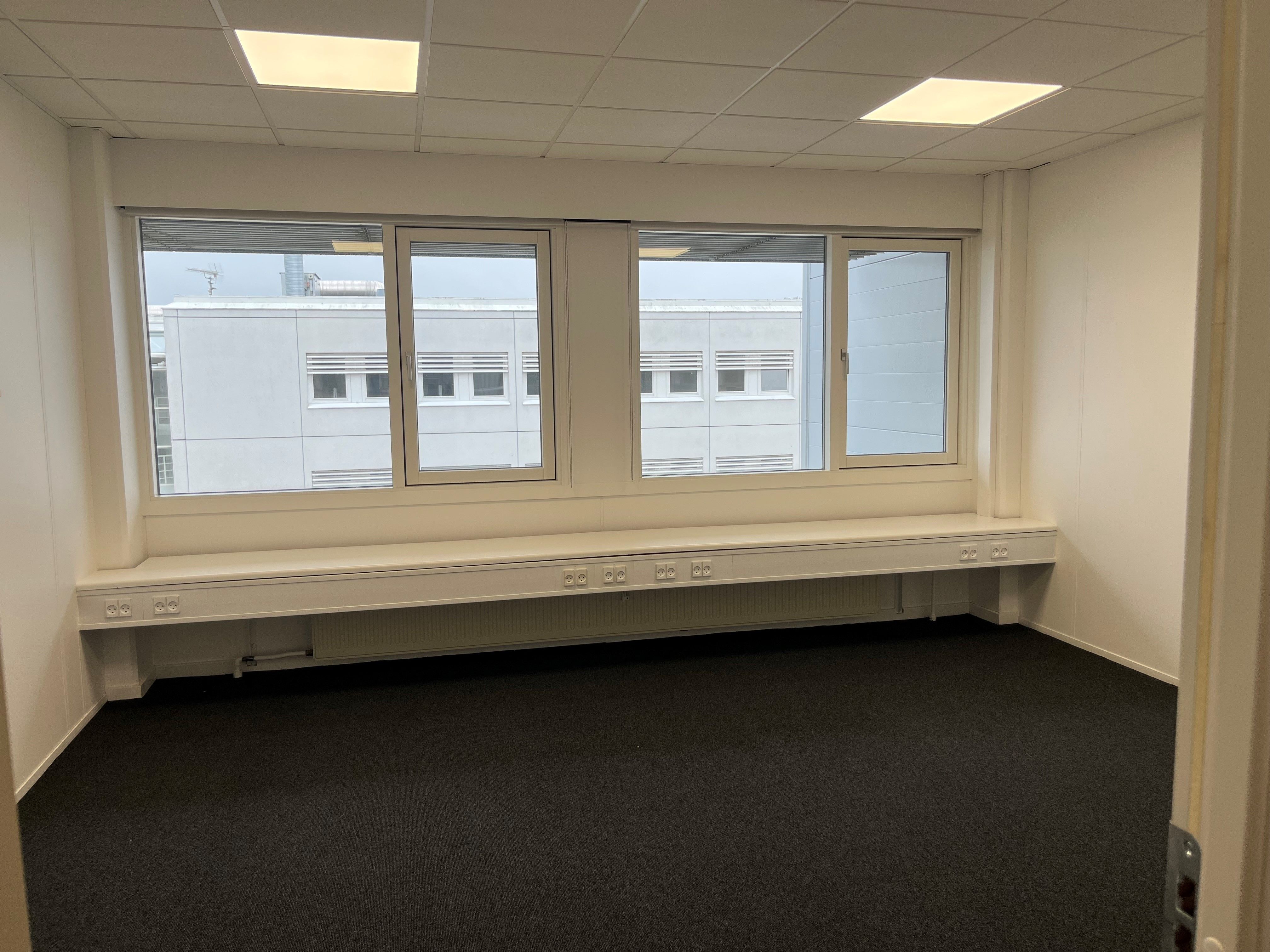 Ledig kontor med tre store vinduer, lysekrone, radiatorer, mørkt gulvtæppe, tre dobbeltstik og en enkeltstik foran vinduet