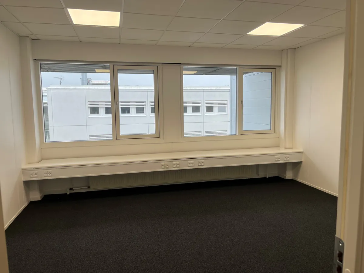 Ledig kontor med tre store vinduer, lysekrone, radiatorer, mørkt gulvtæppe, tre dobbeltstik og en enkeltstik foran vinduet