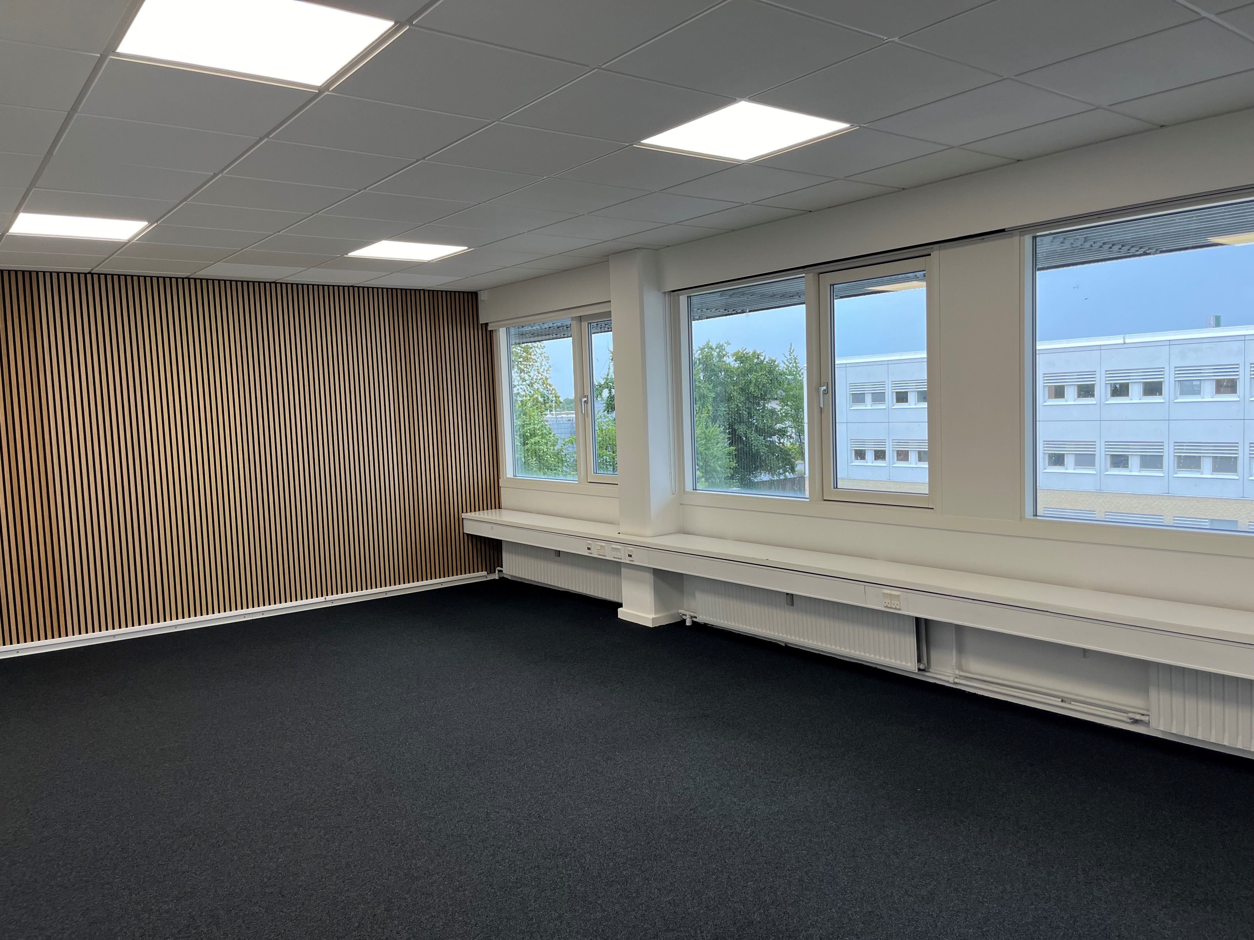 Et stort kontorlokal med lyse loftsplader og høje vinduer, der giver masser af naturlys og udsigt til bygninger og træer