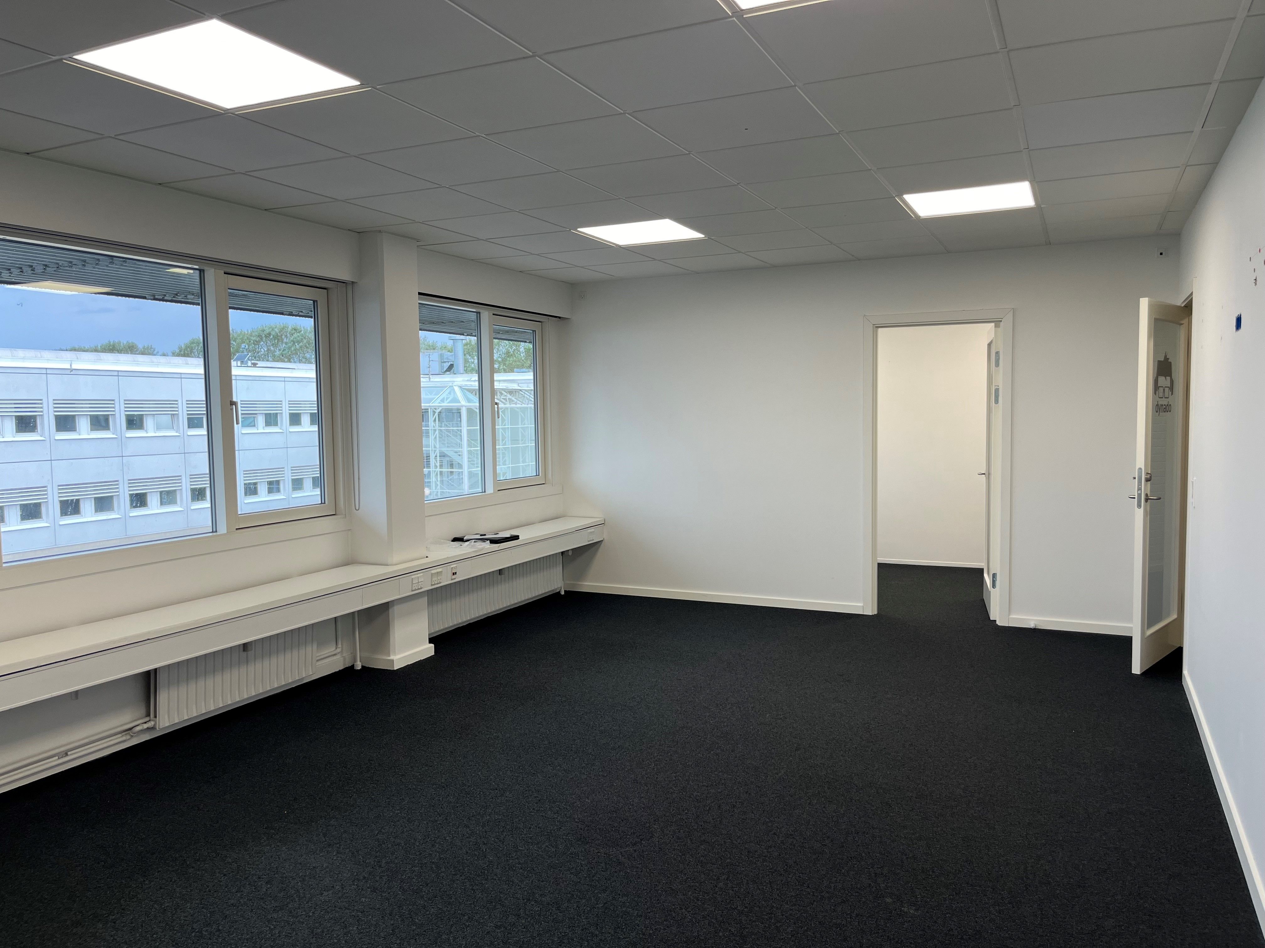 En stor tom kontorrum med hvide vægge, højt gulv og hvide loftspaneler med indbygget belysning samt vinduer og døre
