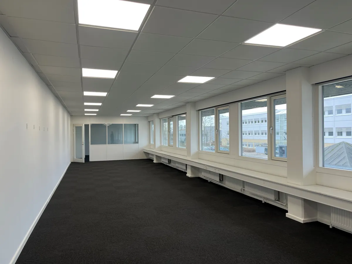 Kort gang med lyse loftslamper og gulvtæppe, langs vinduer med lysegardiner og hvide bænke, der er knyttet til varmeapparater