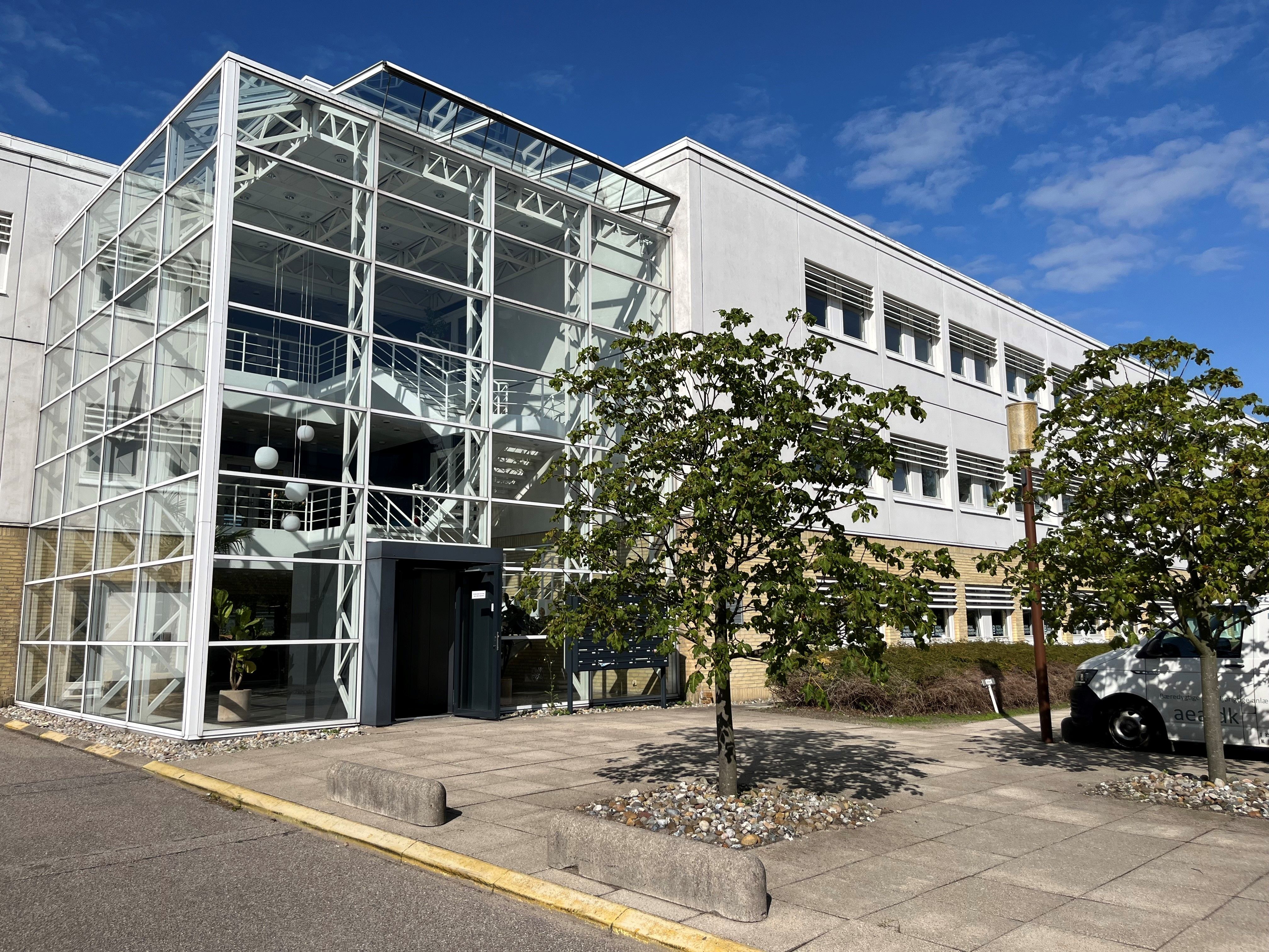 Moderne kontorbygning med stort glasparti og glasdækket trappe. Pæne grønne omgivelser med træer og buske. En hvid køretøj