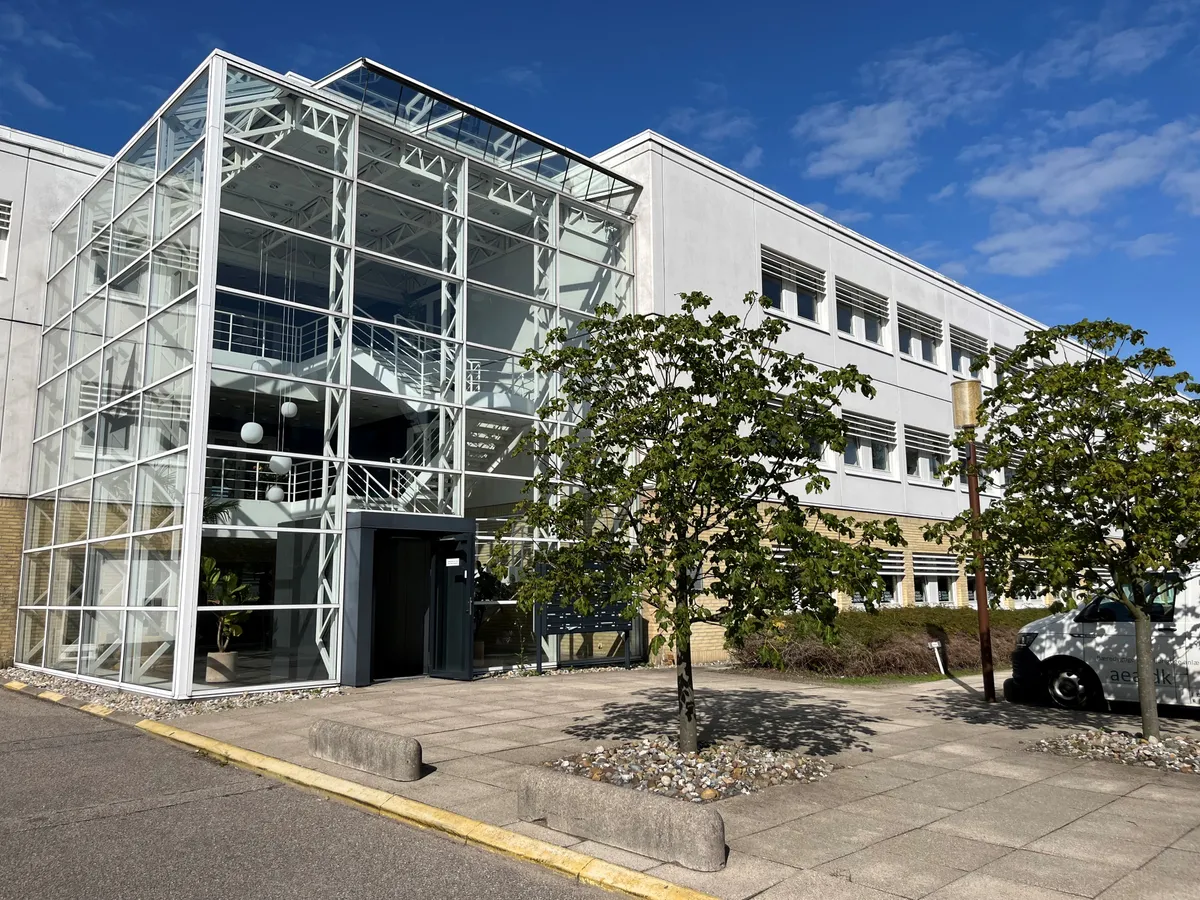 Moderne kontorbygning med stort glasparti og glasdækket trappe. Pæne grønne omgivelser med træer og buske. En hvid køretøj