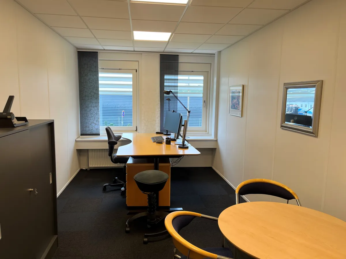 Kontorrum med skrivebord, computer, stol, lampe, kabinet, radiator og to rundere borde med stole i moderne kontorfællesskab