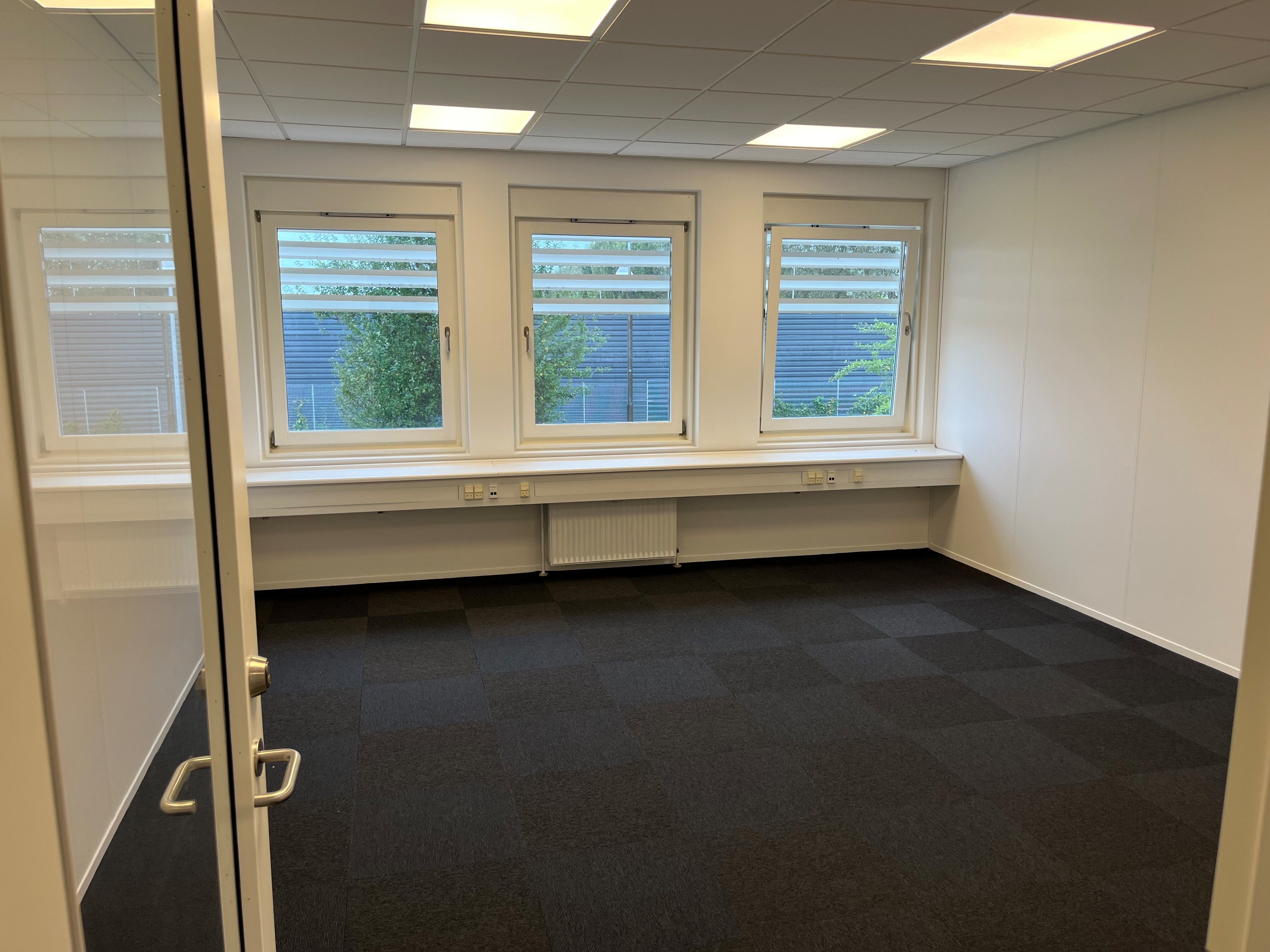 En tom kontorrum med tre store vinduer, skarpe hvide vægge, mørke gulvbelægning og to radiatorer under vinduer