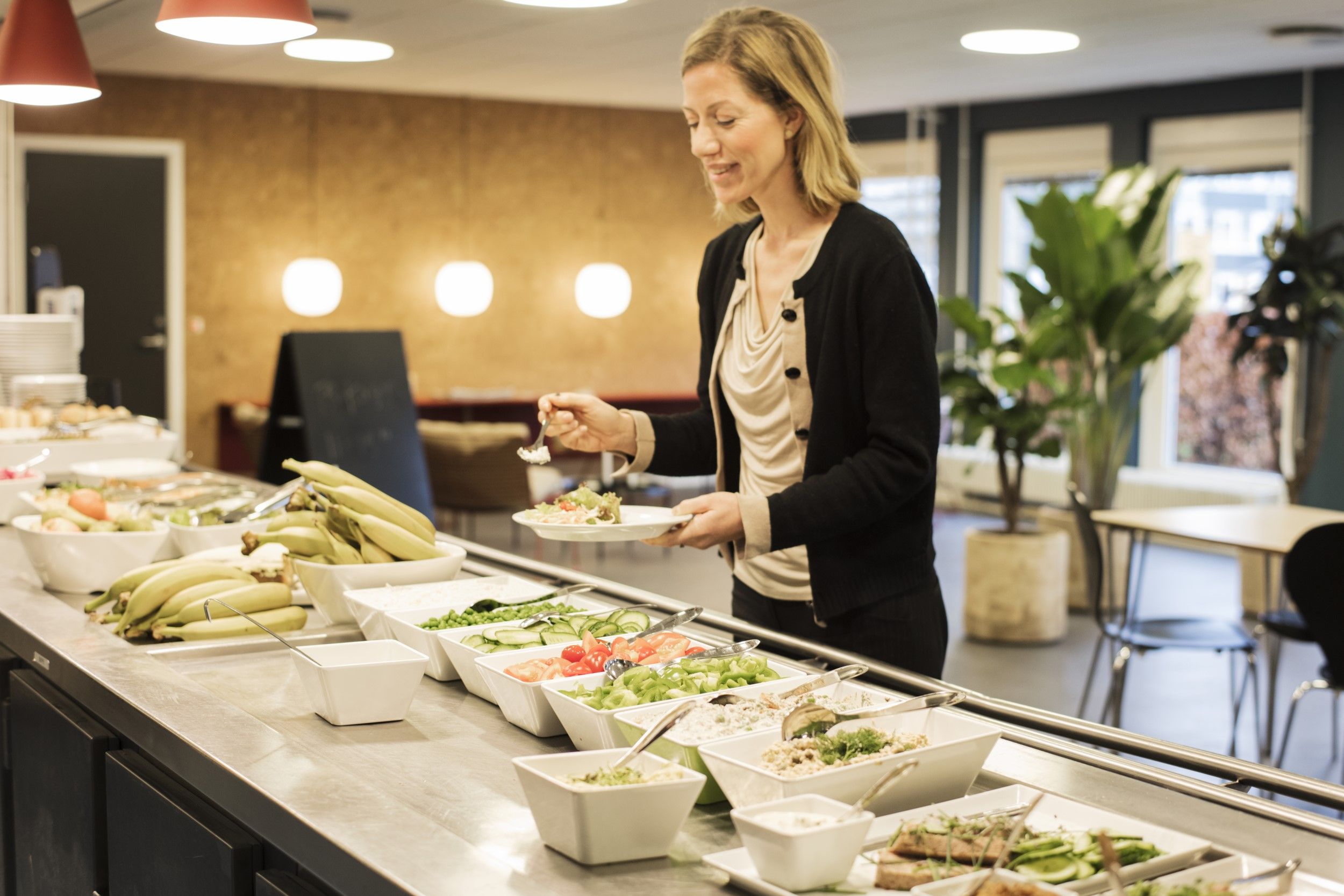 En kvinde serverer en salat i en moderne kantine med grønne planter og lyse loftslampe