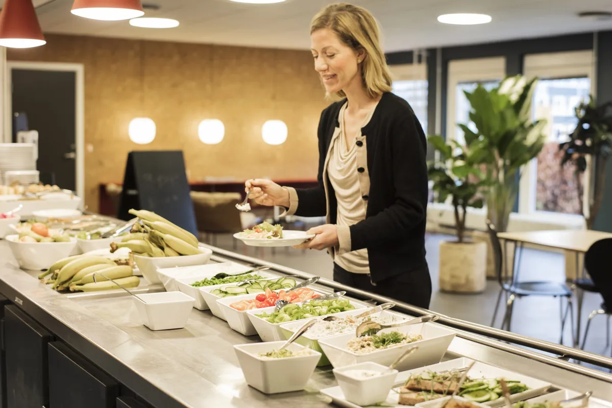 En kvinde serverer en salat i en moderne kantine med grønne planter og lyse loftslampe