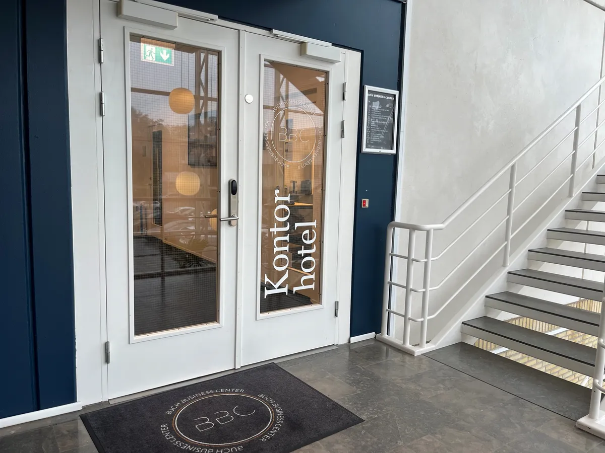 En hvid indgangsdør til Kontorhotellet i Buch Business Center med glasdøre og et glasvindue med refleksioner af en gade og