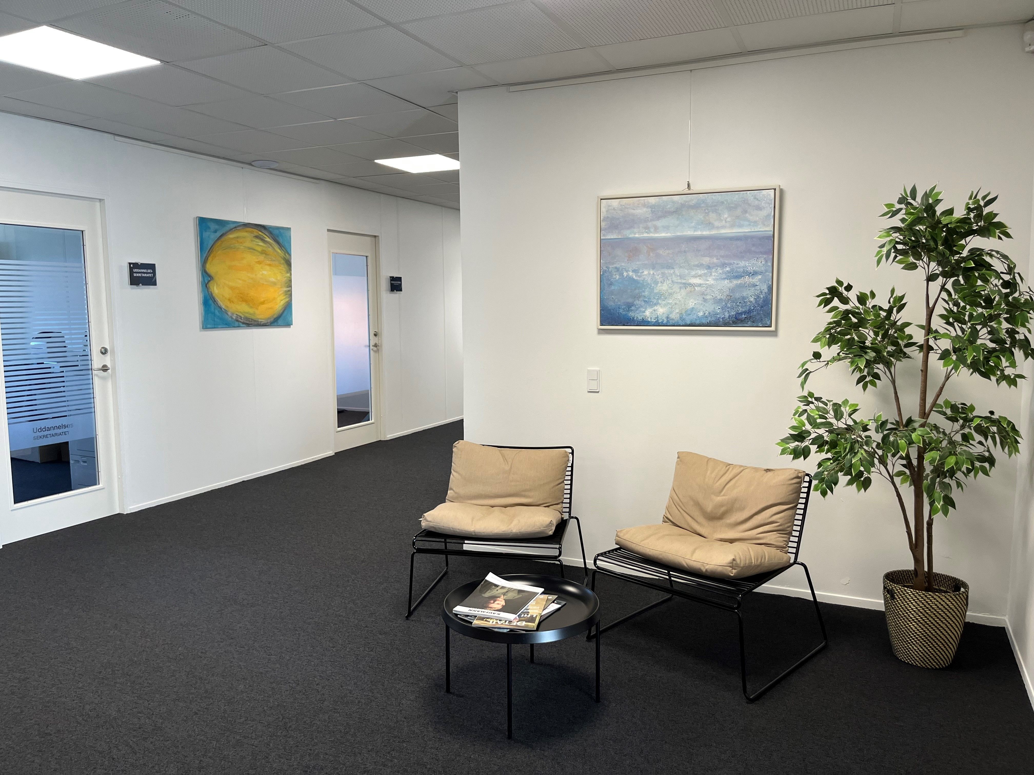 To lænestol med puder står ved en bord og en plante i en rummelig lobby med moderne indretning og kunstværker på væggen