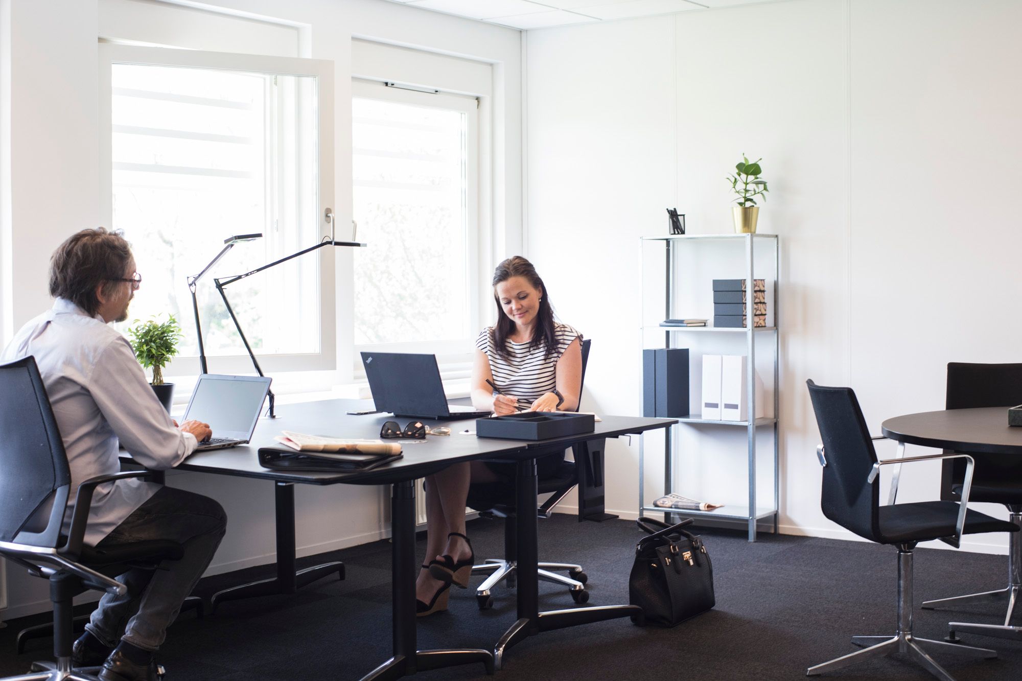 To personer sidder i et moderne kontor med to laptops og en stol med høj ryglæn foran et bord med et skab og en plante