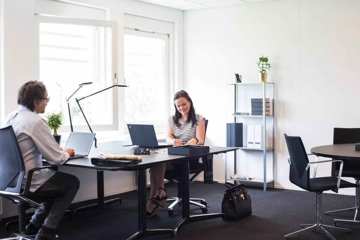 To personer sidder i et moderne kontor med to laptops og en stol med høj ryglæn foran et bord med et skab og en plante