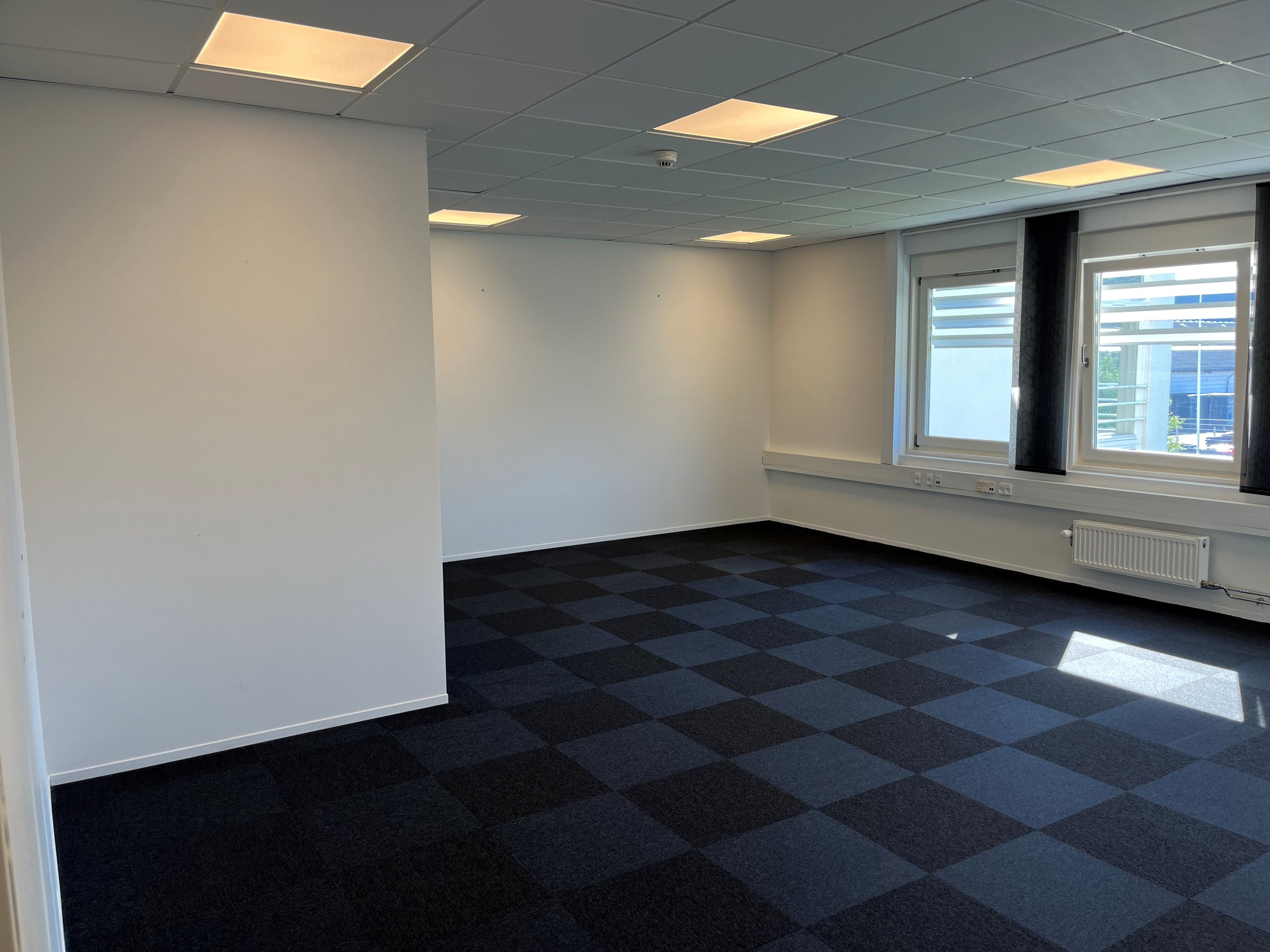 En tom kontorlokale med hvide vægge og loft samt en tæppegulv med skakfigurer og smukke vinduer med sollys