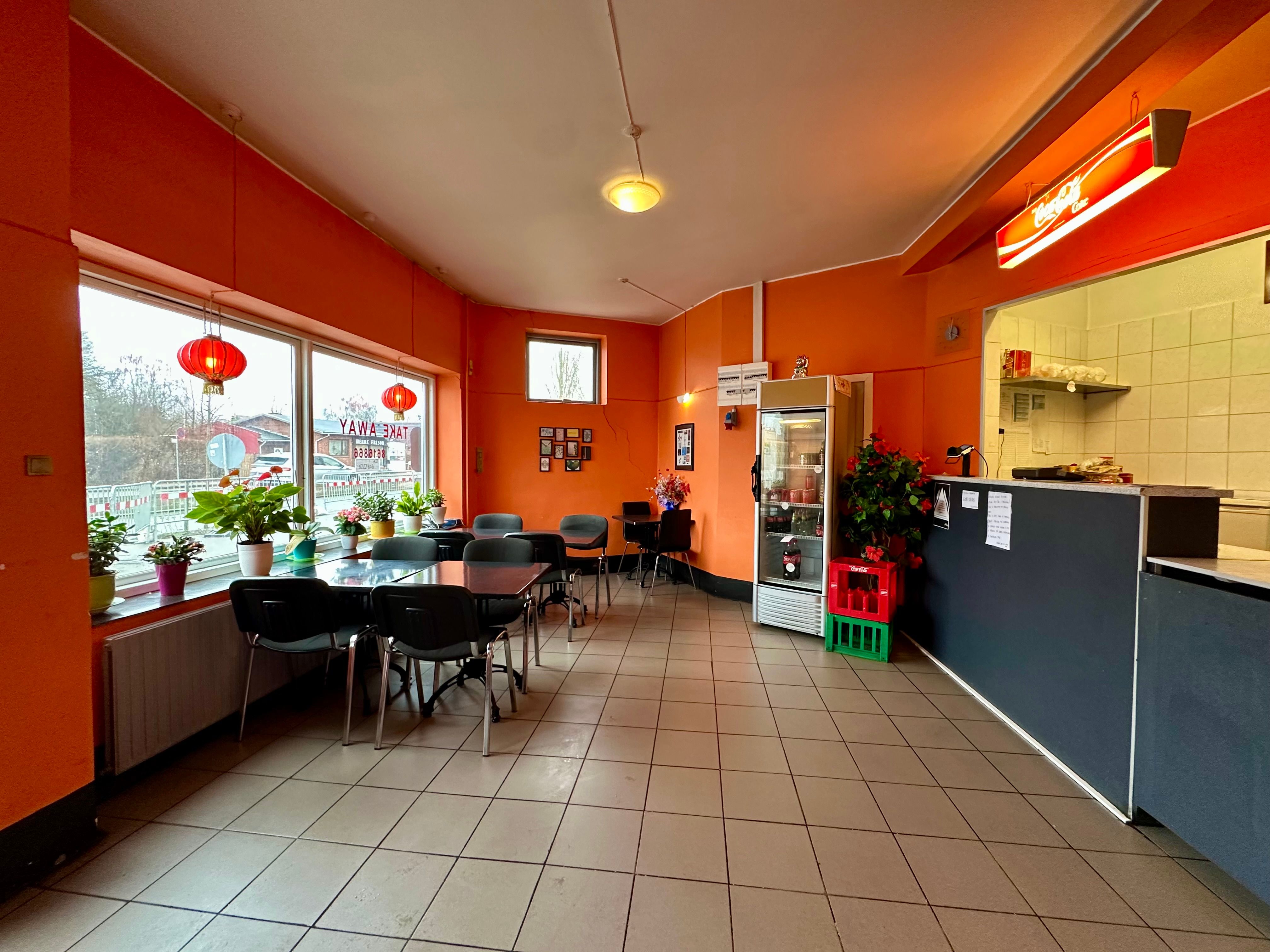 En spisehal med borde og stole, lysekroner, plantepoteter, glasdøre og en Coca-Cola-reklame.