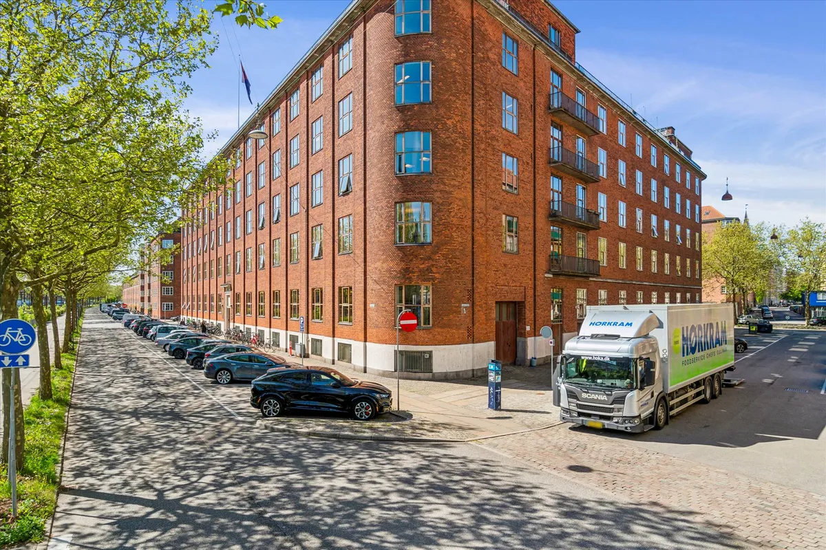 En stor bygning med mange vinduer og balkoner. En stor lastbil står parkeret foran bygningen. Der er flere biler parkeret