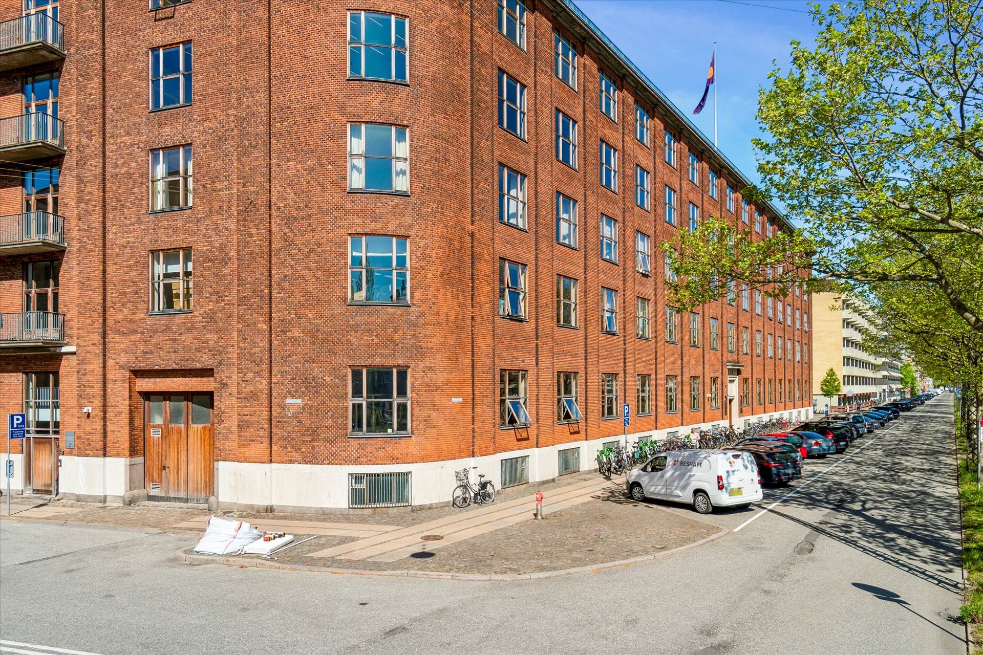 Stort rødt murstenshus med flere etaager og mange vinduer står på en bred gade med parkerede biler og cykler langs siden