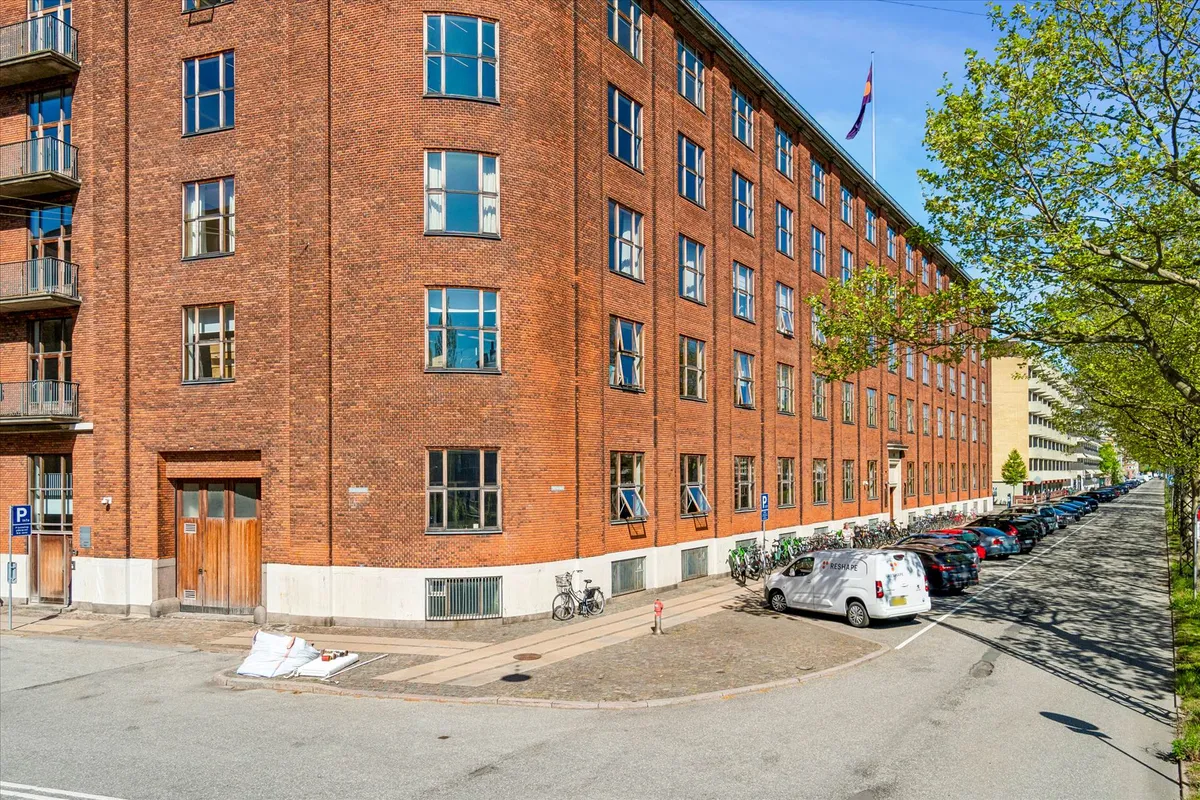 Stort rødt murstenshus med flere etaager og mange vinduer står på en bred gade med parkerede biler og cykler langs siden
