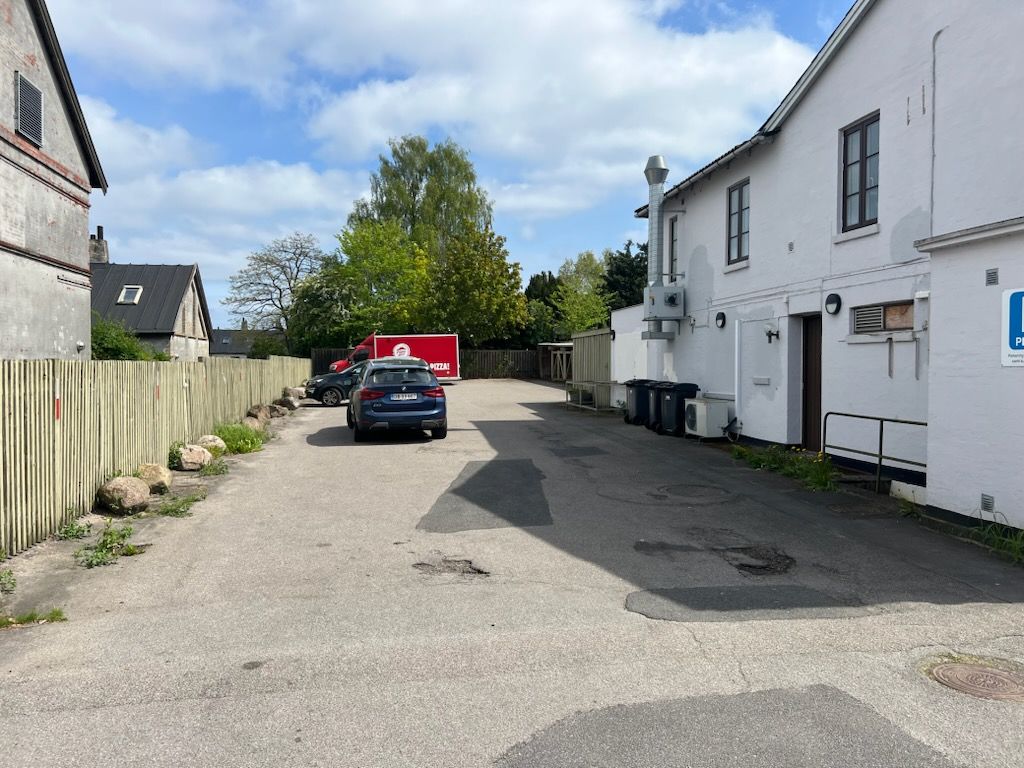 Privat parkeringsplads med lyseblå bil parkeret og rød leveringsvogn i baggrunden, sammen med en hvid bygning og røde