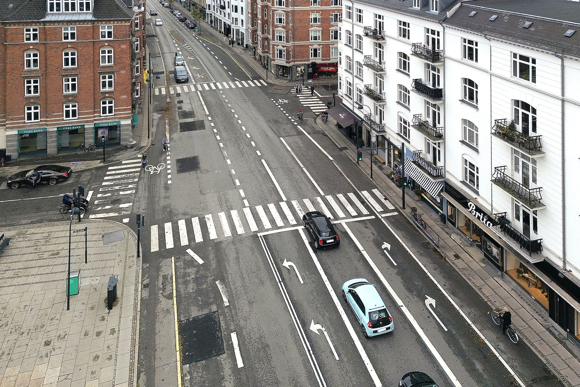 To biler kører på Strandvejen i Hellerup, og folk krydser gaden ved en fodgængerovergang. Bygninger med røde og hvide