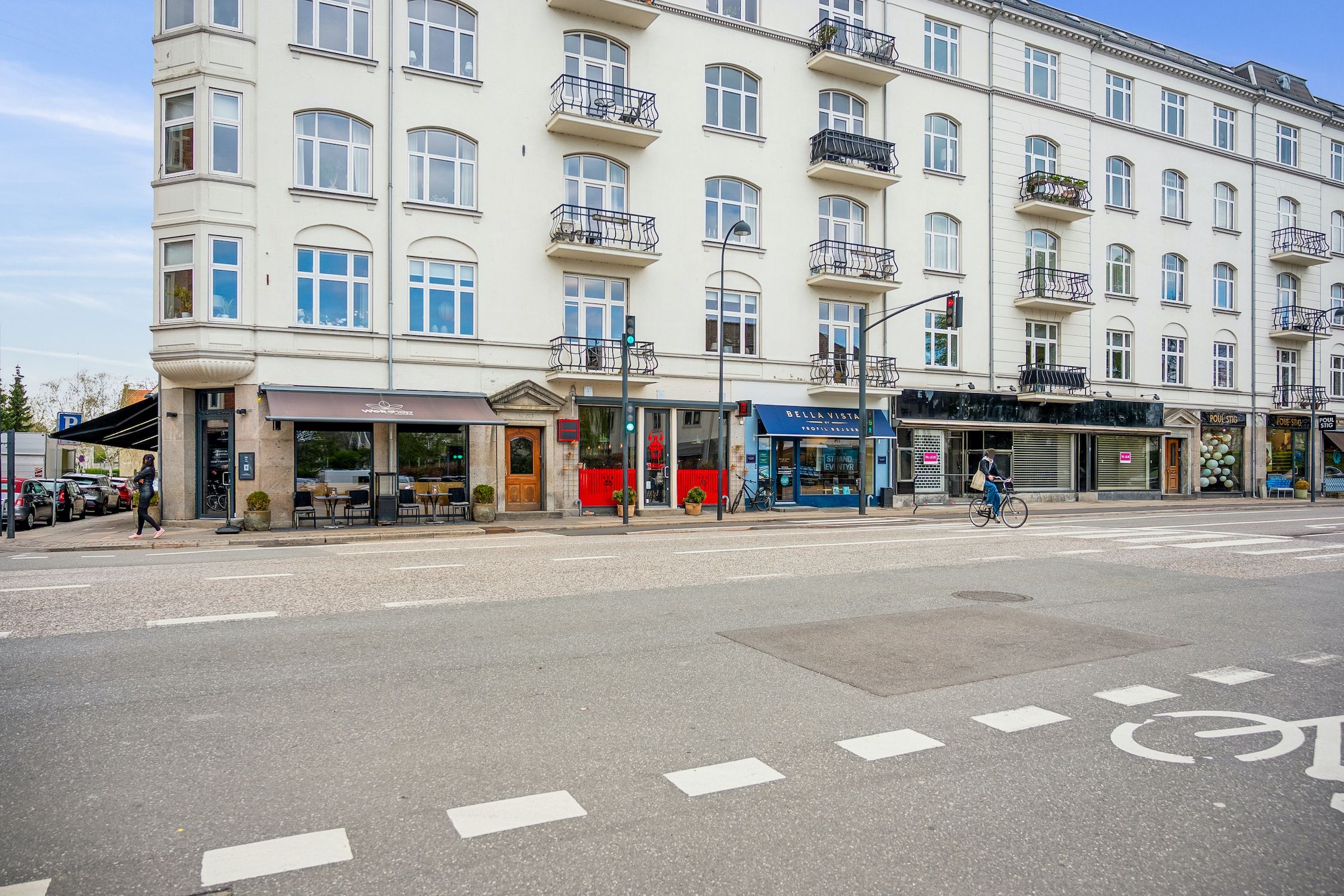 En person cykler gennem et tomt vejafsnit, mens en anden person går på fortovet uden for en stor bygning med mange