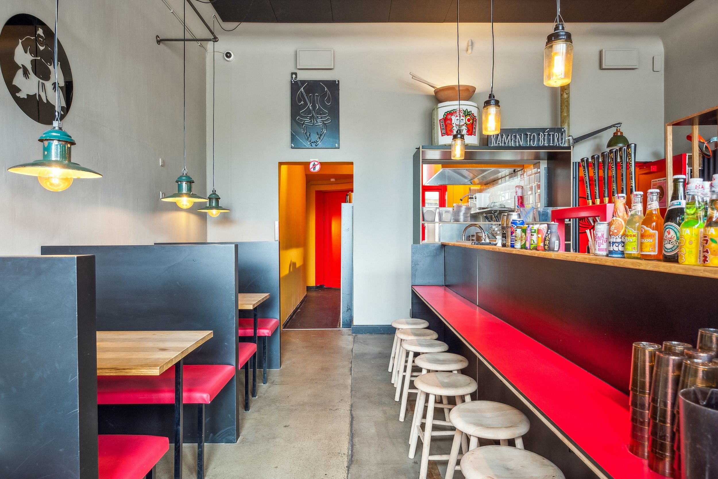 Lyst restaurant med loftslamper og en bar med soda, øl og ølglas. Sorte sofaer med røde sæder og træborde.