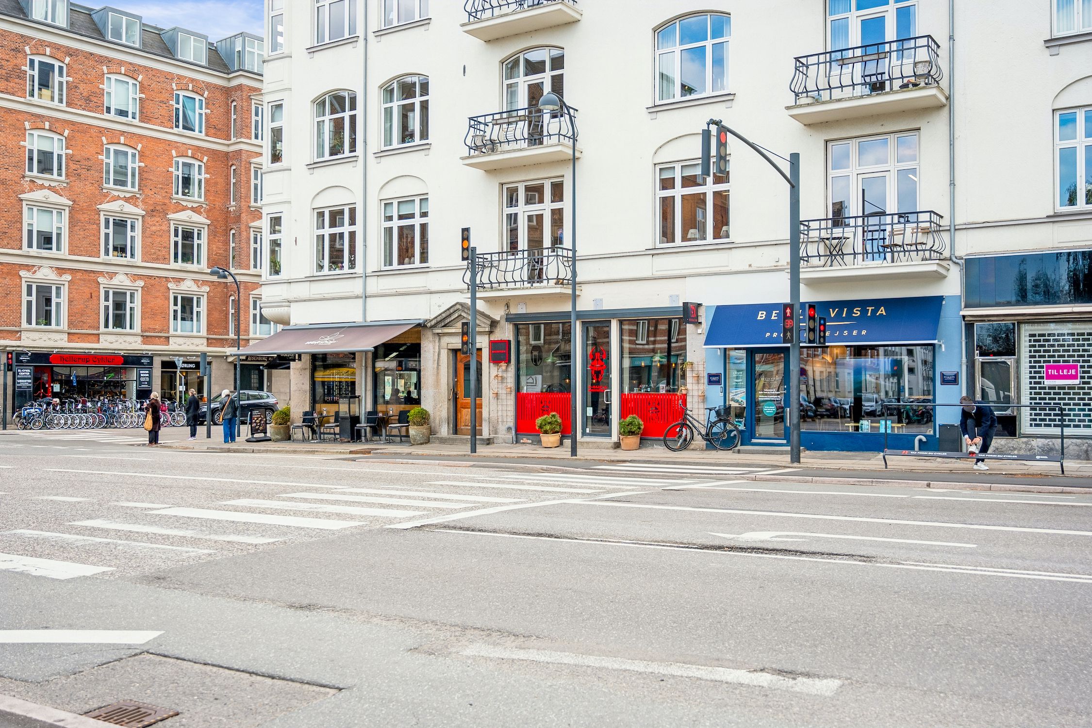 Solrige gader med moderne bygninger, gadebelys og terrasser. Gågå og cykelparkering med mennesker og planter. Butikker med