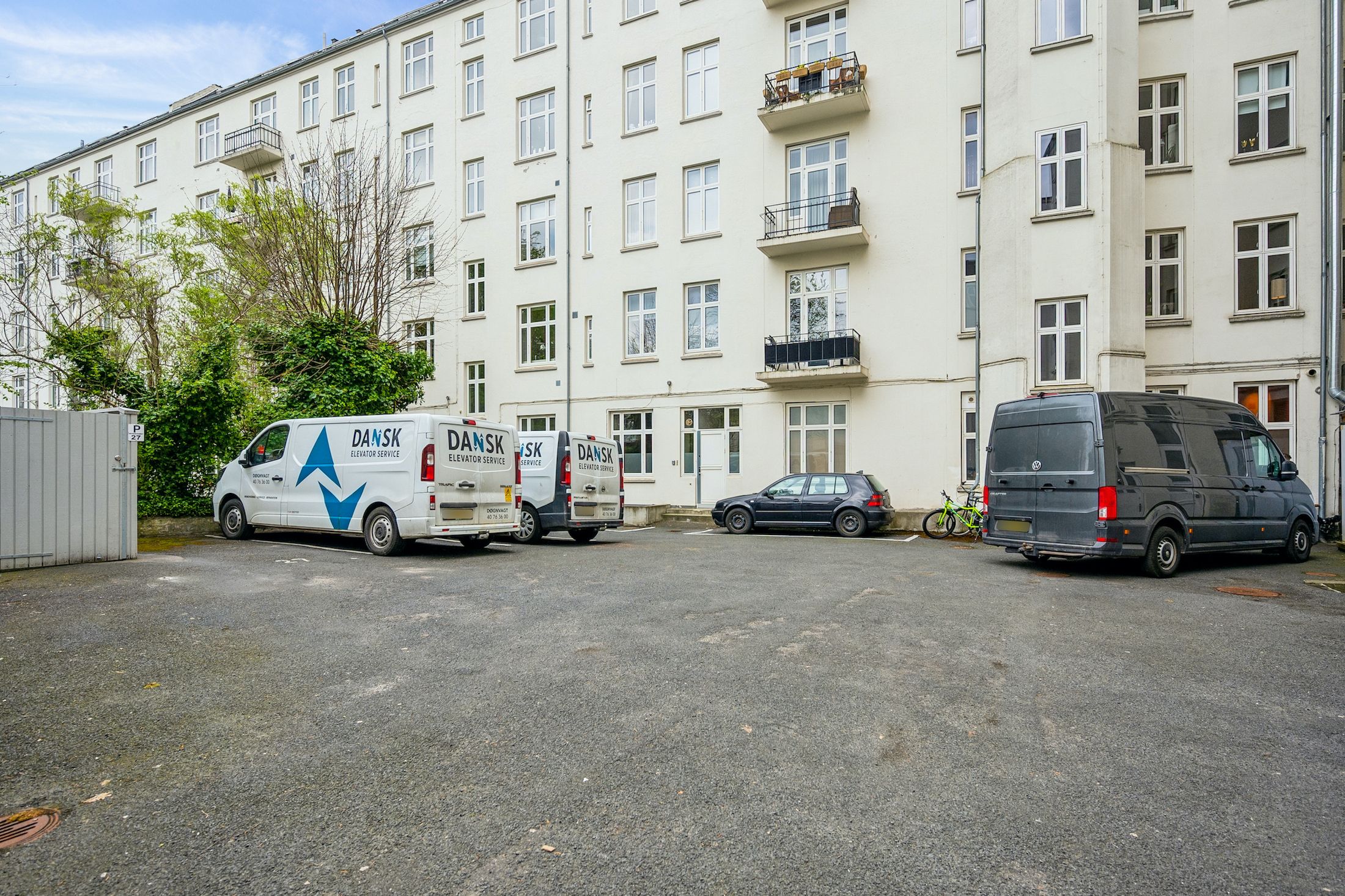 Foran et lejlighedshus parkerer to servicevogn, en Dansk Elevator Service, og en Volkswagen. To cykler står parkeret ved