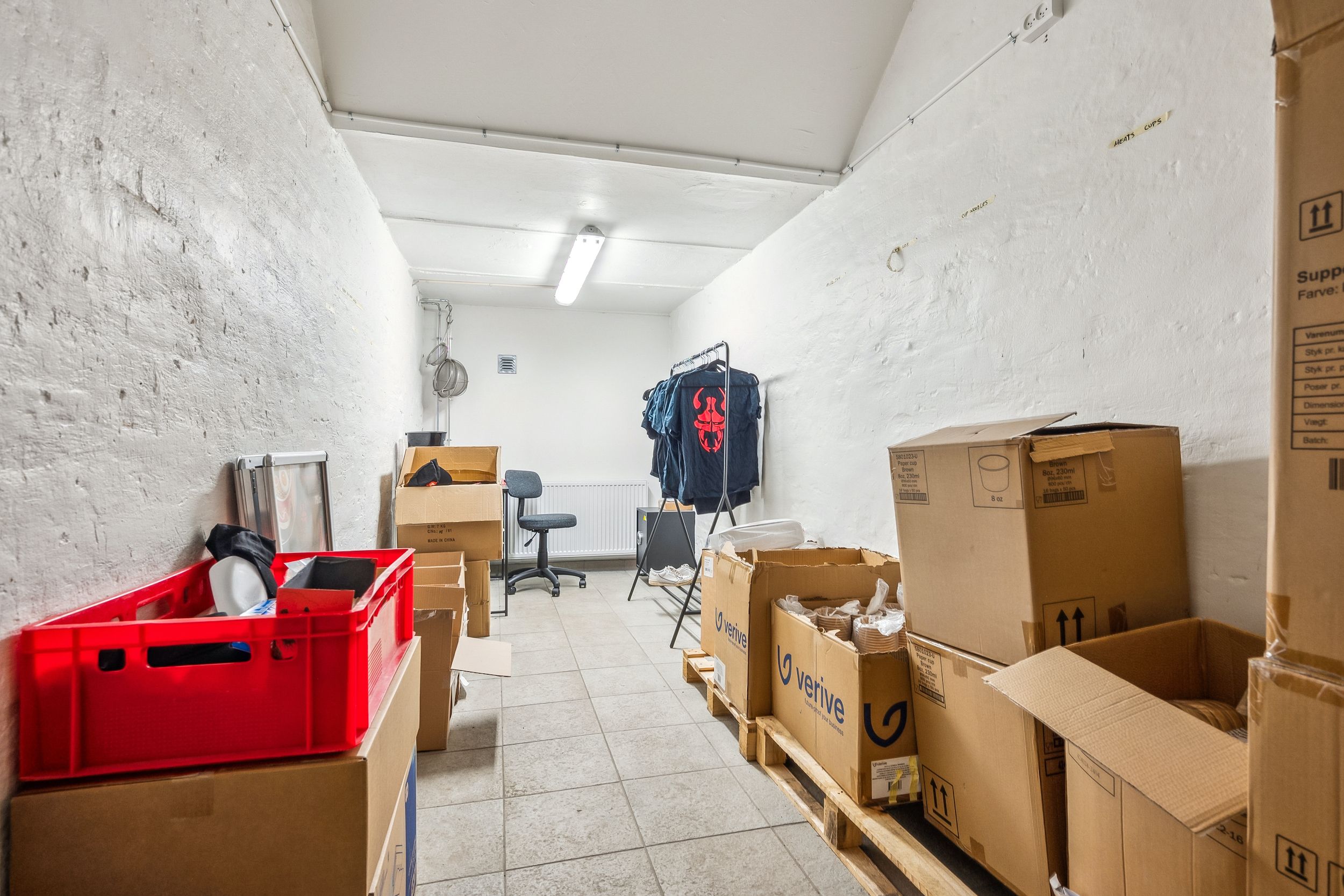 Kasser og pakker på lagerplads med lysegul belysning og hvidt vægmateriale, og tøj hængende på klædestander.