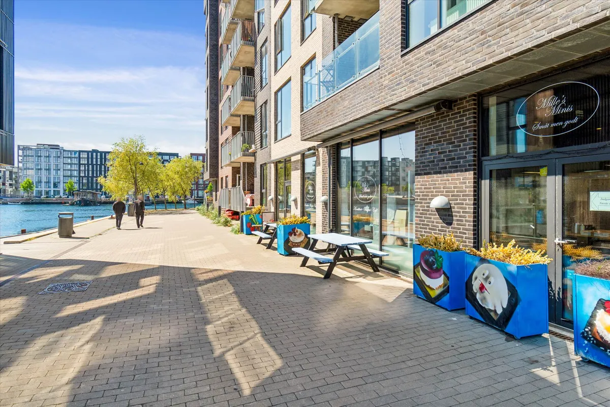 En moderne bygning med udsigt til vandet og grønne træer, med en butiksforretning og udendørs spisesteder med udsigt til