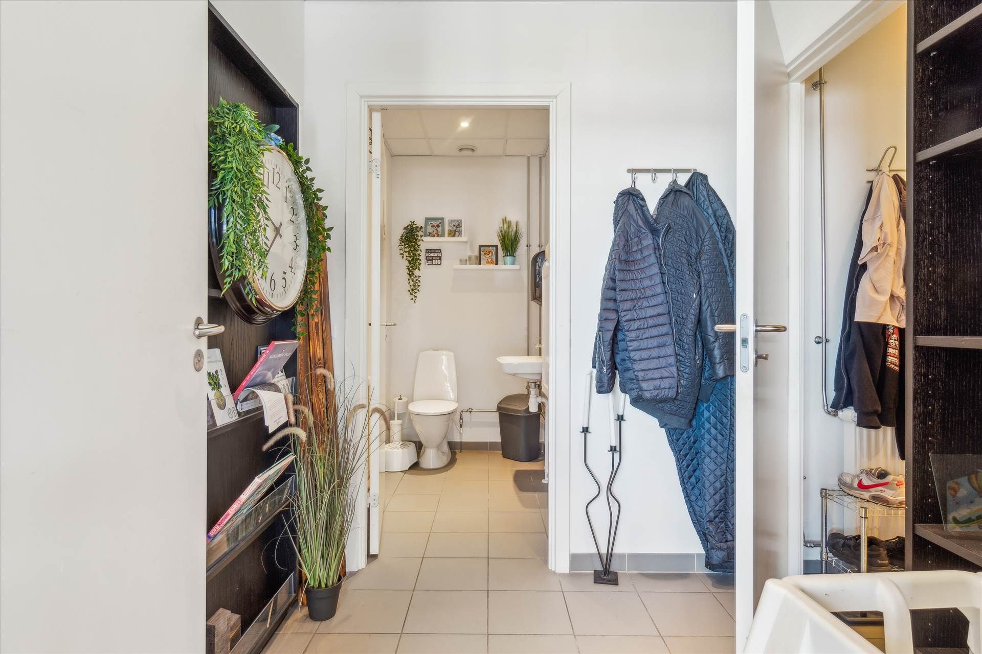 En moderne toiletrum med toilet, håndvask og toiletpapir samt garderobe med tøj og sko samt et kældre med ur og plante