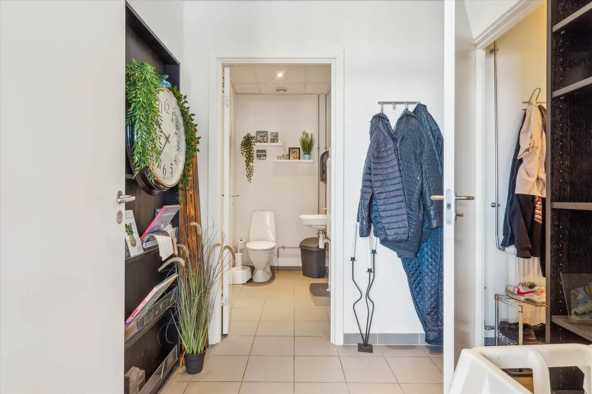 En moderne toiletrum med toilet, håndvask og toiletpapir samt garderobe med tøj og sko samt et kældre med ur og plante