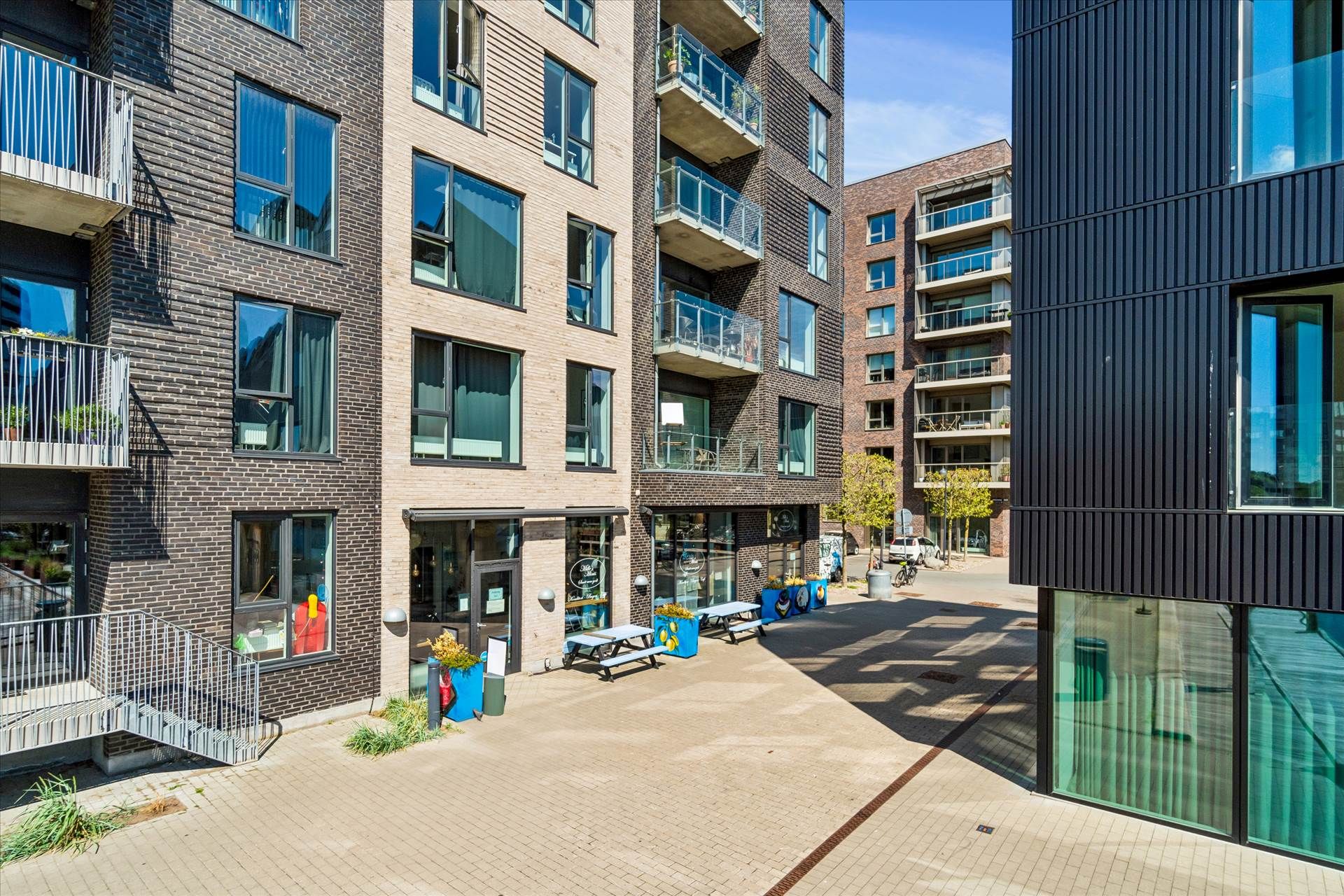 Moderne bygninger med terrasser og balkoner langs en gågade. En række små haver og grønne vægplantede potter. Et moderne