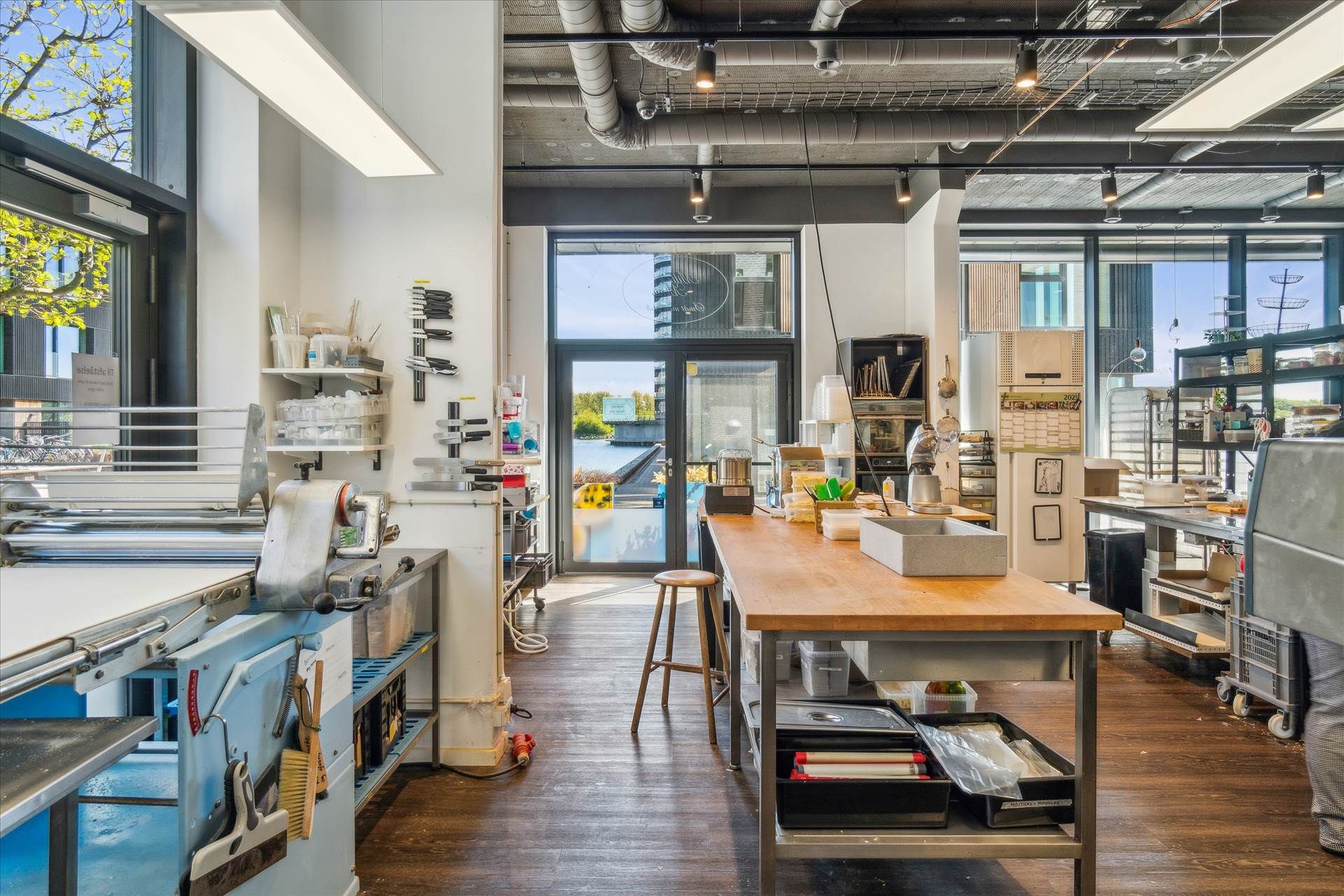 En stor, åben arbejdsplads med vinduer og lyse loftslamper, flere værktøjer og maskiner, et stort træbord og en høj stol