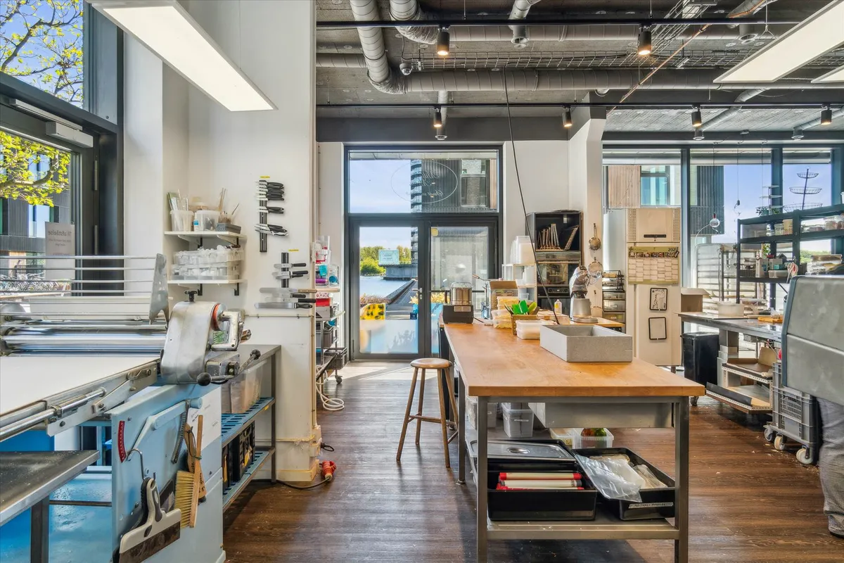 En stor, åben arbejdsplads med vinduer og lyse loftslamper, flere værktøjer og maskiner, et stort træbord og en høj stol