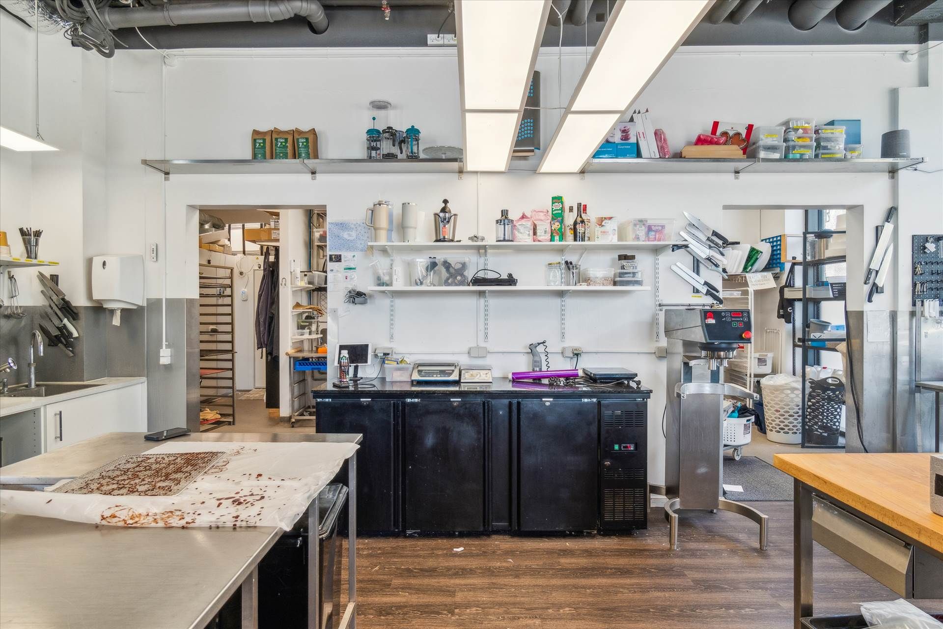 Moderne køkken med flere apparater og strygebrætter, trægulv, hvide vægge og lyse loftslampe samt skarpe kantede møbler