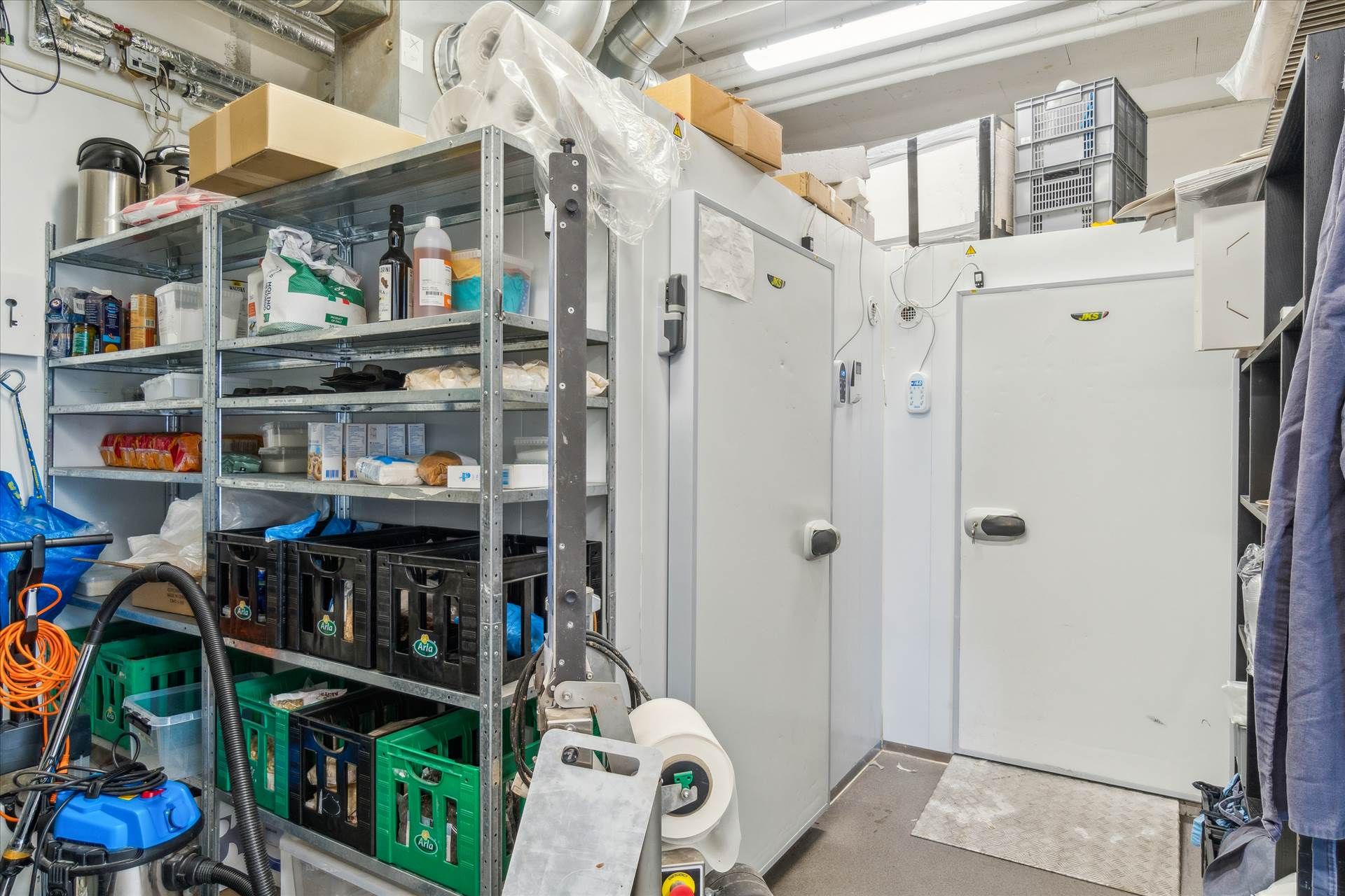 En opbevaringsrum med flere hylder, to køleskabe, kasser, bøtter, en støvsuger og diverse ting og apparater.