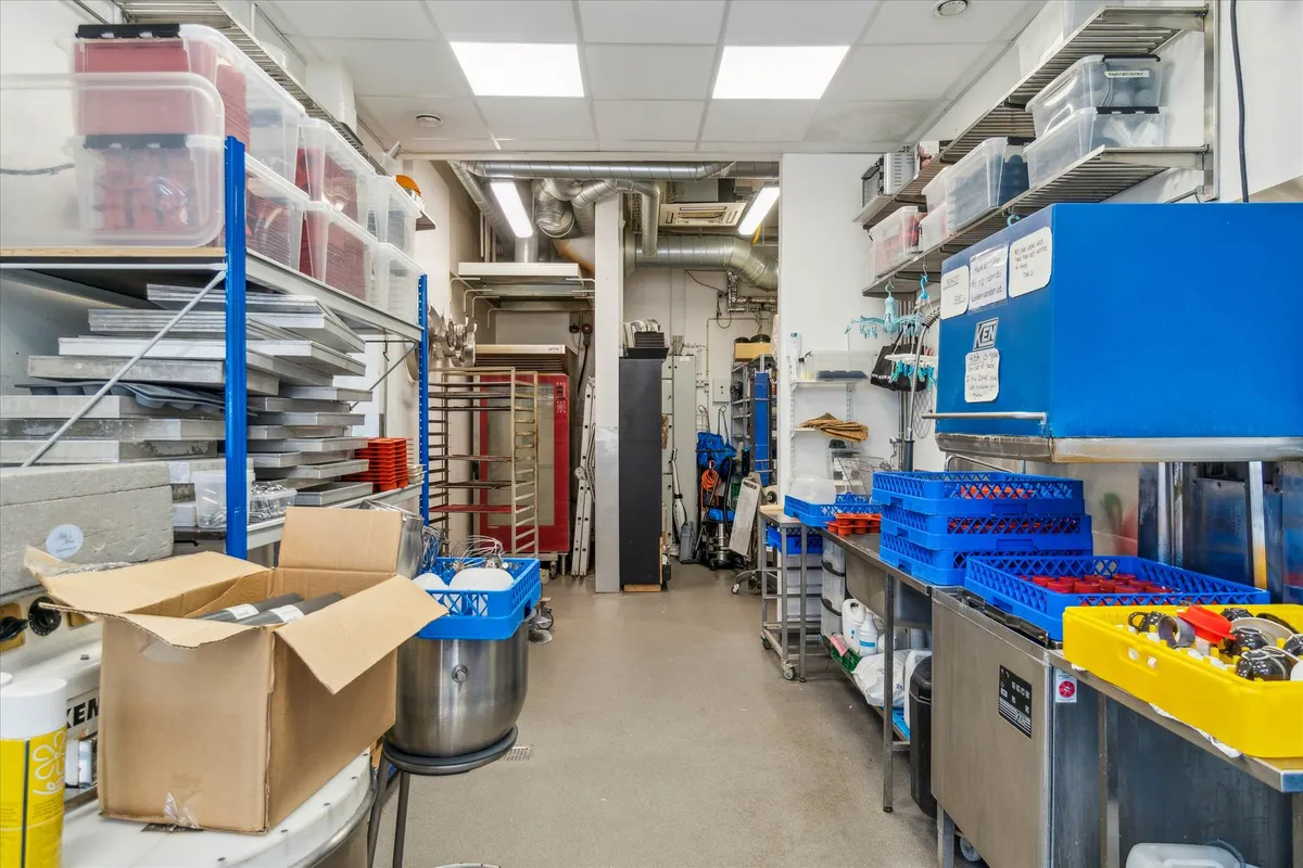 Et stort lager med mange hylder og beholdere, blå kassere og rullekurve, en stor metalbøtte, røde hylder og en rød ovn, og